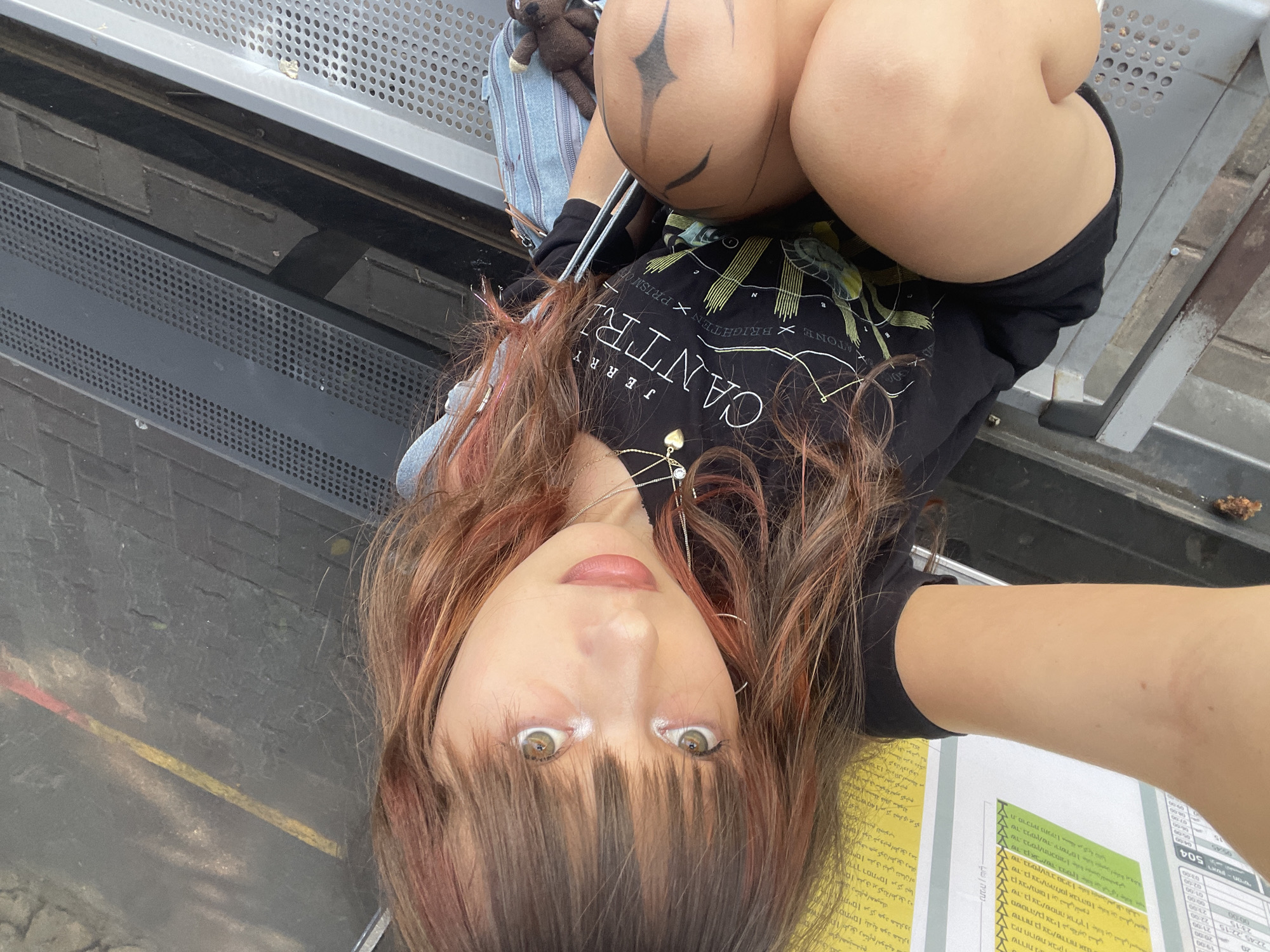 A young woman with long, reddish-brown hair with lighter streaks sits on a metal bench, her body angled towards the camera. She is wearing a black t-shirt with a graphic print and black shorts. She has a light skin tone, with visible eye makeup and lip gloss. A silver necklace with a heart pendant is visible around her neck. Her right leg is bent, and her left arm is extended out of frame. A denim bag with a small brown stuffed animal is resting on the bench beside her. The bench appears to be in an outdoor public space, with a textured ground visible in the background. A partially visible yellow and red striped marking is on the ground. Printed materials with Hebrew text are on the left side of the image. The overall impression is a casual, self-portrait style photograph.
