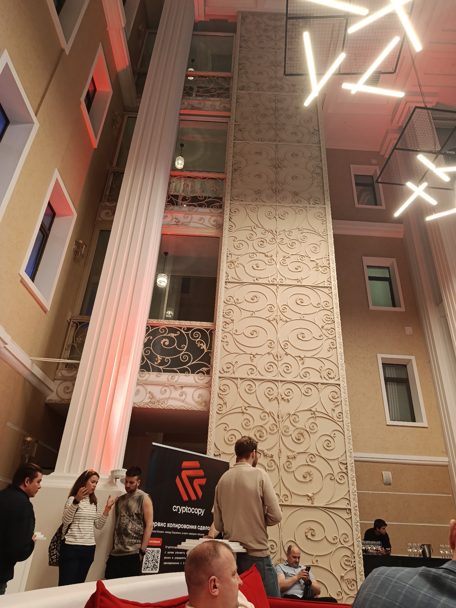 The image shows an indoor event in a building with a multi-story atrium. The architecture is ornate, featuring decorative columns, window detailing, and a patterned wall element. Lighting fixtures of contemporary design are visible overhead.
Several people are present. Some are gathered near a banner with the word "cryptocopy" and some Russian text, suggesting a crypto-related event. Others are sitting or standing nearby, engaging in conversation or looking at their phones.
The location is possibly in Moscow, Russia, based on the Cyrillic text on the banner.