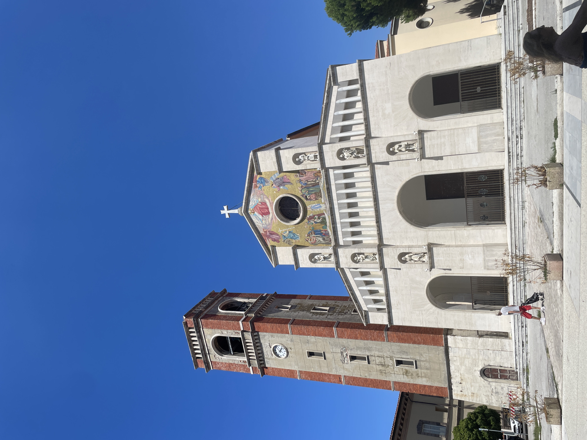 The image depicts the exterior of the Duomo di Viareggio, officially the Chiesa di San Giovanni Bosco, located in Viareggio, Italy. The facade is prominent, with a mosaic above the main entrance depicting religious figures under a white cross. To the left, a tall, brick and stone bell tower with a clock face stands adjacent to the church. The sky is clear and blue. In the foreground, a person wearing red shorts and a white t-shirt walks with a black wheeled bag. Several planters with dry vegetation are present on the paved area in front of the church. The architecture suggests a Mediterranean style.