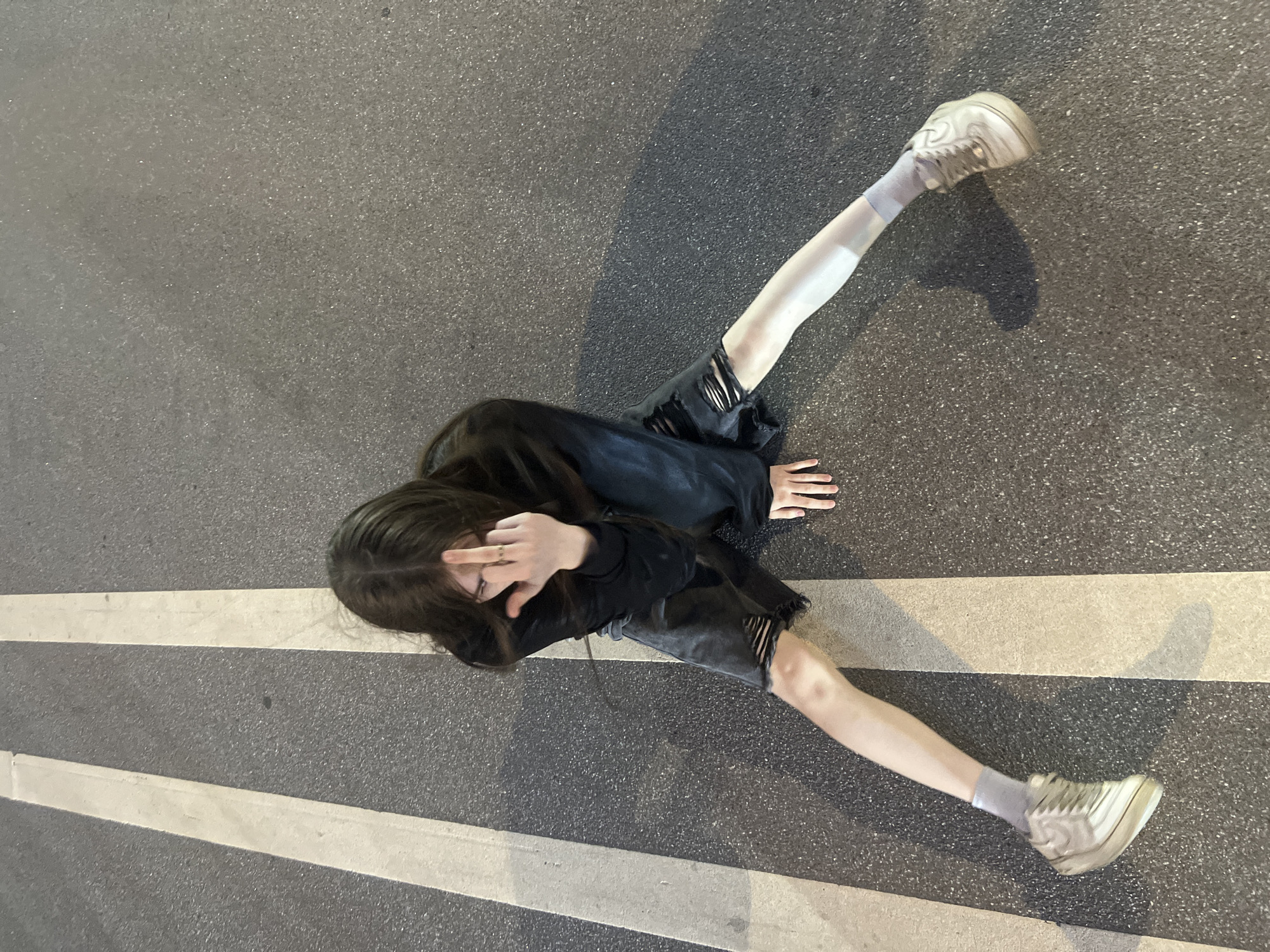 The image displays an individual seated on an asphalt road surface under low-light conditions, consistent with an urban setting in Saint Petersburg, Russia. The individual has long dark hair and is wearing a black long-sleeved top, dark ripped shorts, light-colored socks, and light-colored sneakers. Their legs are spread wide apart, with one leg extended across one of two parallel white painted lines on the asphalt. The individual's left hand is placed on the ground, while the right hand is raised, extending the middle finger towards the camera. A ring is visible on the middle finger. Shadows are cast on the dark grey, granular asphalt surface from an unseen light source.