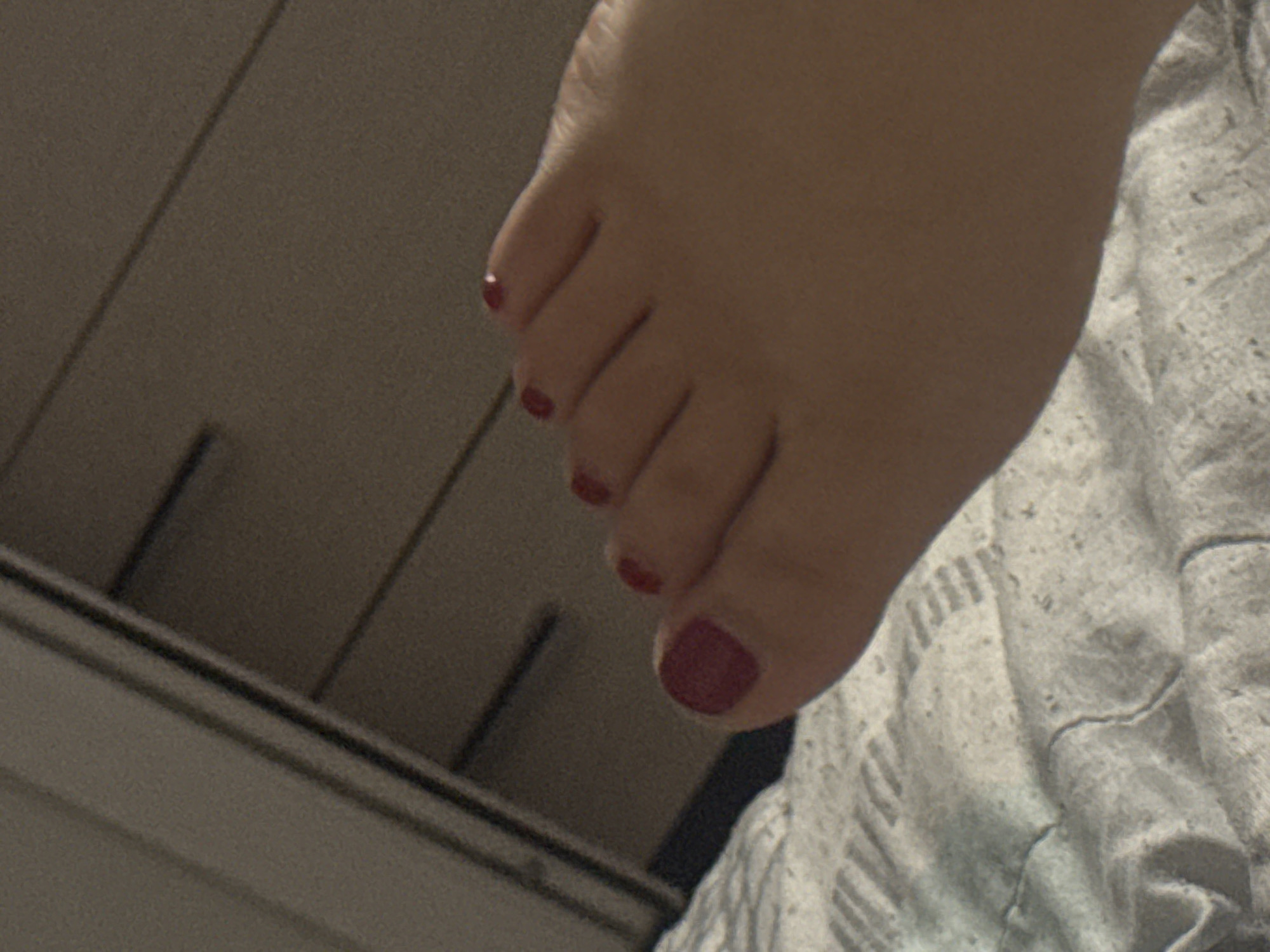 The image is a close-up, low-angle shot of a person's left foot with dark red nail polish on the toenails, resting on a white, textured blanket. The background consists of dark brown wooden drawers with horizontal lines, suggesting furniture. The lighting is dim. The location in Saratov, Russia cannot be determined from the visual information provided.