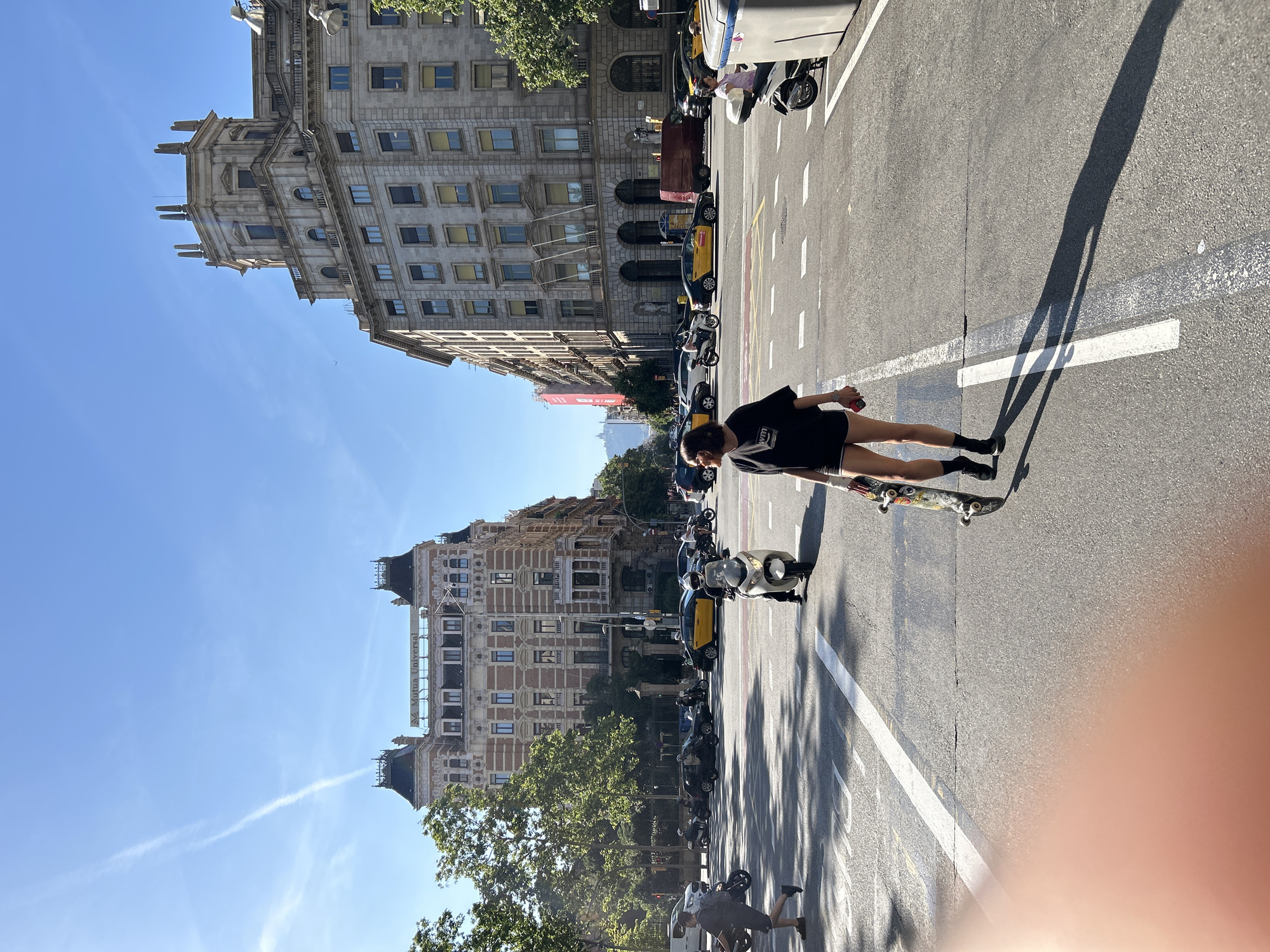 The image depicts a street scene in Barcelona, Spain. The photo is taken at street level, oriented along the asphalt roadway. A person wearing a dark shirt and black socks is standing on a skateboard in the street. Yellow taxis and scooters are parked along the sides of the street, and buildings rise on either side. The sky is blue. The street is marked with white lines indicating lanes. The Mutua Universal building is visible in the background. The image seems to be partially obscured in the lower right-hand corner.
