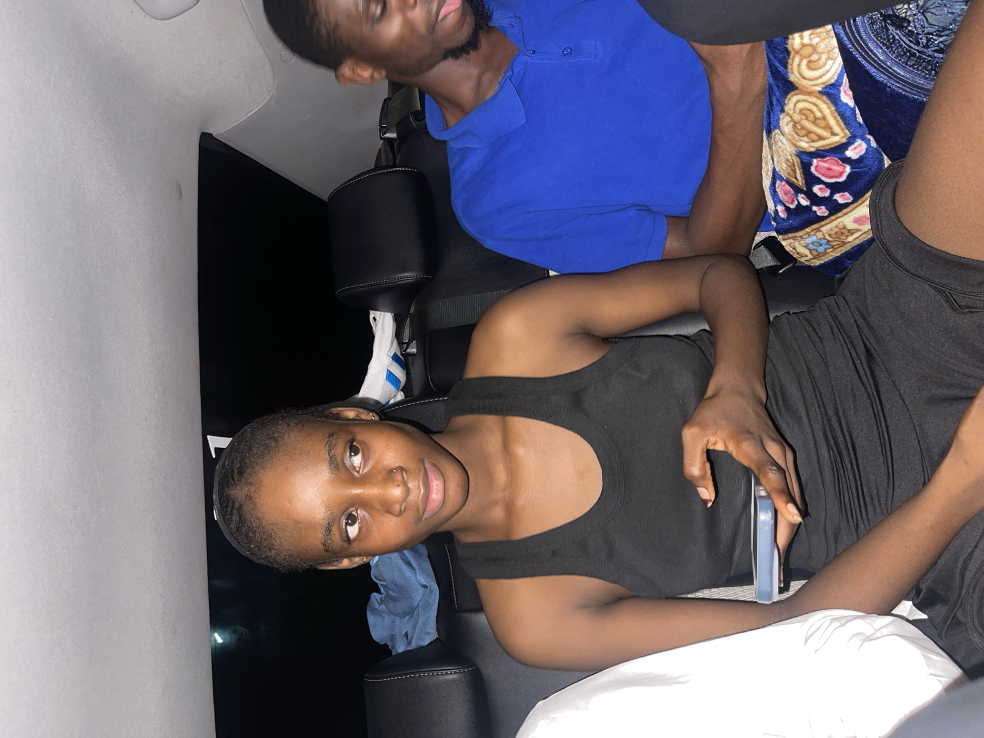 The image depicts a group of young people inside a car, likely a sedan, with a view of the interior. The scene appears to be at night, with the interior lights of the car illuminated and the outside dark. The seating arrangement suggests they are in the back seat, with the front passenger seat and headrests of the car visible.
In the foreground, a young woman is the primary subject, positioned on the left side of the frame. She is wearing a black tank top and black shorts, and her hair is pulled back. She is looking to her right, and her right hand is holding a light blue smartphone.
To her right, a young man is seated, wearing a bright blue polo shirt. He is looking down and to his left, with his tongue slightly protruding from his mouth. He has short dark hair and a beard.
The car interior is visible, including black leather seats with white stitching, seat belts, and headrests. A folded blue cloth is on the seat behind the woman. On the man's lap is a folded blanket with a colorful pattern of blue, white, pink, and brown.
The overall lighting suggests an indoor setting at night, consistent with being inside a vehicle in an urban environment. While the image does not contain specific landmarks to definitively place it in Abuja, Nigeria, the context of a car interior at night is common in urban settings worldwide.