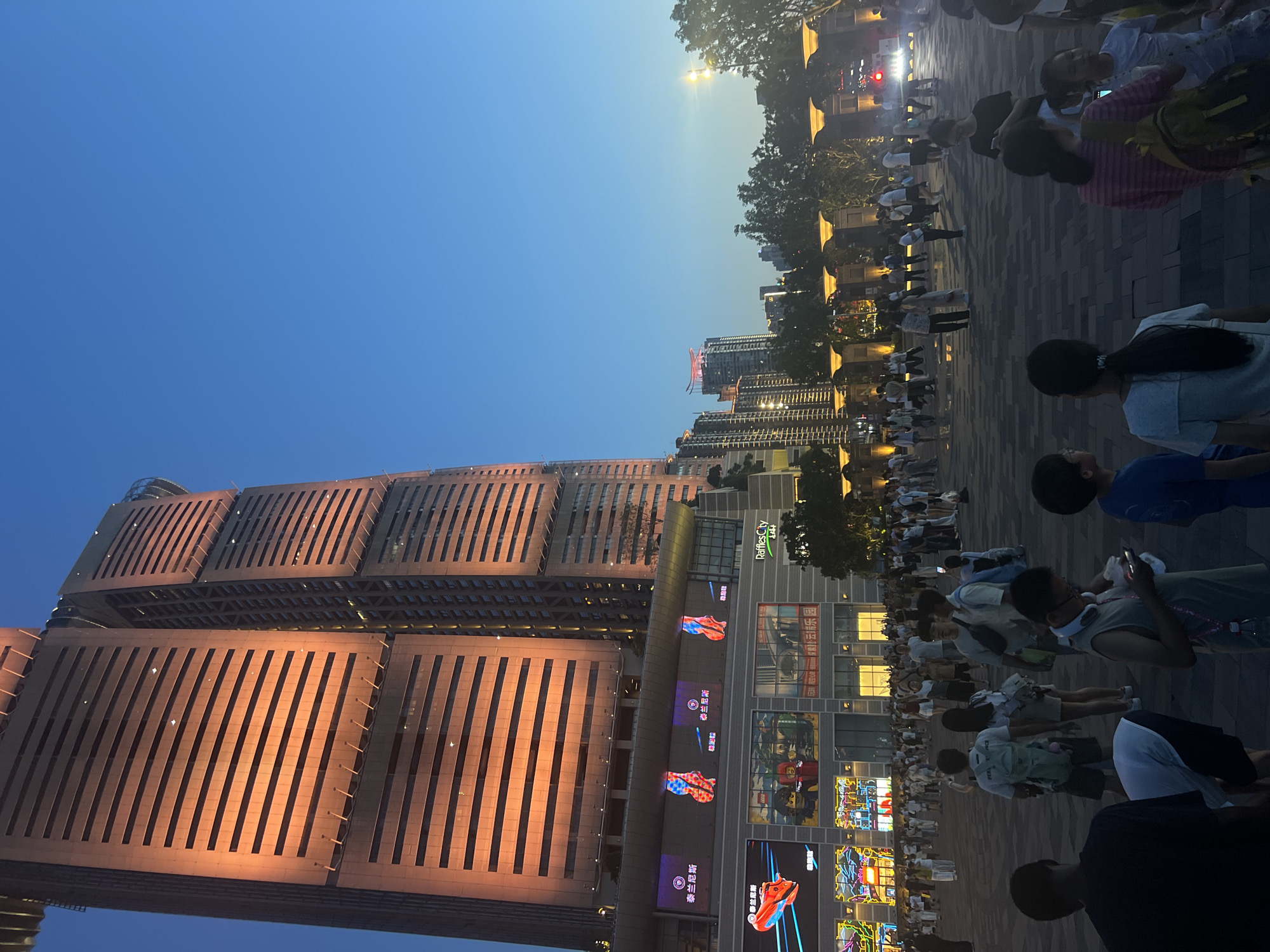 The image depicts an evening scene in front of a modern, illuminated skyscraper in Yuzhong District, China. The skyscraper, which appears to be the Raffles City Chongqing complex, dominates the background with its warm orange lighting. Below the skyscraper, a multi-level shopping mall features brightly lit advertisements, including images of shoes and Lego figures. A large crowd of people, dressed in casual attire, are gathered in the plaza area in front of the mall. Some individuals are standing, some are walking, and a few appear to be looking at their phones. The sky is a deep blue, transitioning into twilight. Trees and ornate lampposts are visible along the periphery of the plaza, and a street with traffic lights can be seen in the distance. The overall atmosphere suggests a public gathering space, likely a popular spot for entertainment and shopping.