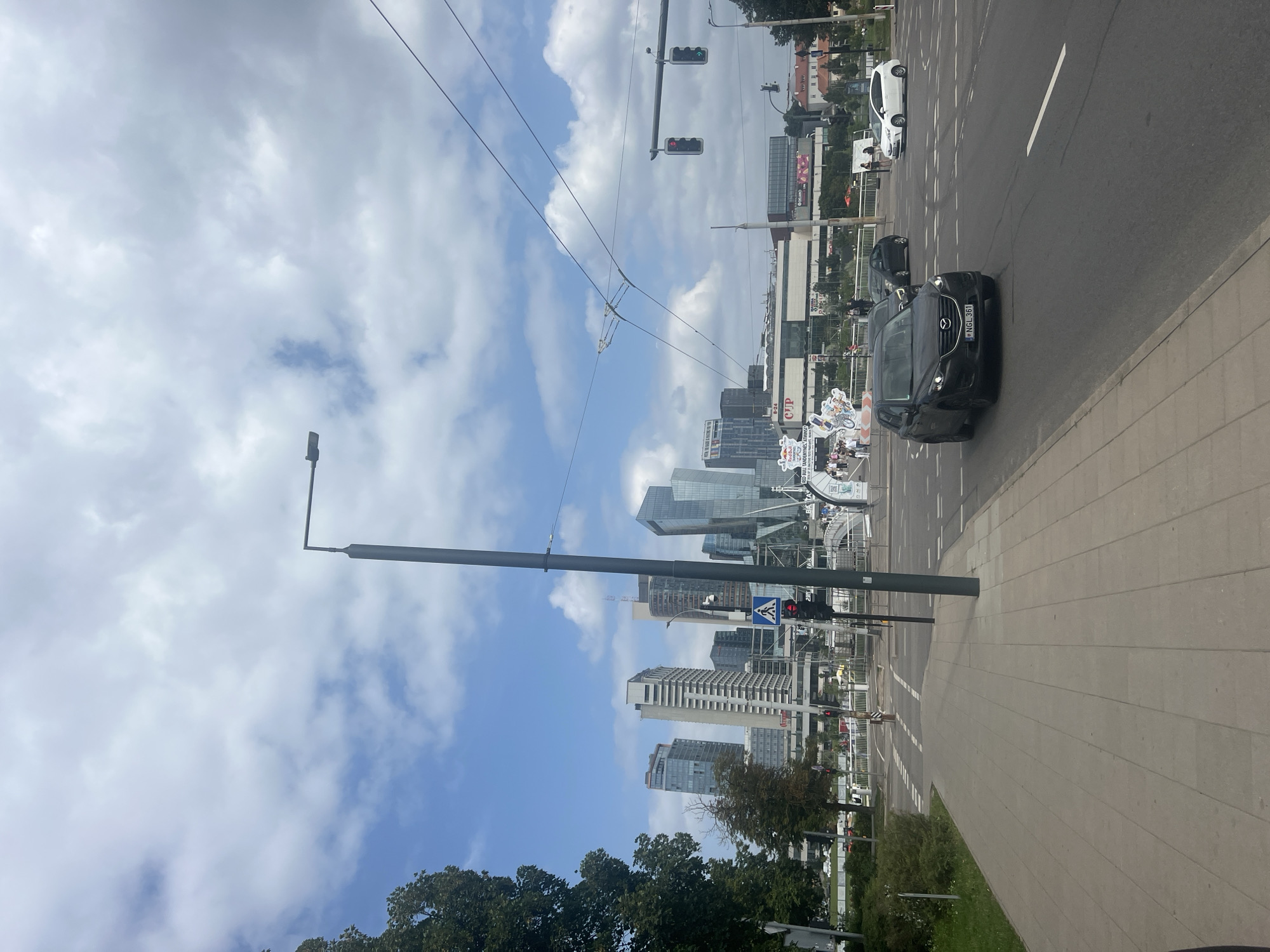 The image captures a street scene in Vilnius, Lithuania, with a view of the modern cityscape in the background. In the foreground, a paved sidewalk runs along a road. A dark-colored Mazda CX-5 is visible on the road, turning right. Several other cars are also present on the road. Traffic lights are visible, with one showing a red light and another showing a green light. A pedestrian crossing is marked on the road. In the distance, several tall buildings and skyscrapers dominate the skyline, characteristic of a business district. Trees and green spaces are present on the left side of the image. Overhead wires and a lamppost are also part of the street furniture. The sky is partly cloudy, with patches of blue visible. A bridge and some event banners are also discernible in the mid-ground, suggesting a possible public event or festival. The overall impression is of a dynamic urban environment on a day with mixed weather.