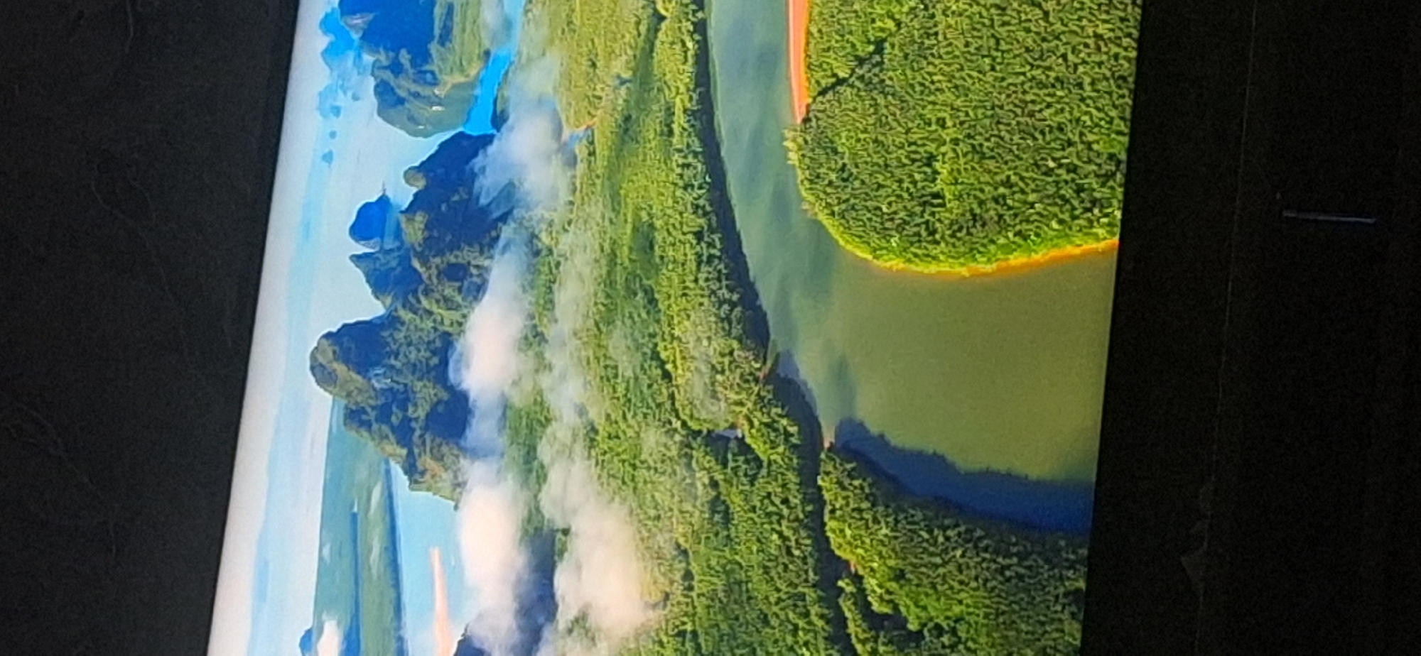 The image displays an aerial view of a coastal landscape with lush green forests, a winding river, and several rocky islands emerging from the water. Wisps of white clouds drift across the scene, partially obscuring some of the islands and the sky. The water is a mix of deep blue and lighter shades, reflecting the sky and surrounding vegetation. The forest canopy is dense, with a vibrant green hue. The overall scene suggests a tropical or subtropical region, likely a national park or a protected natural area. There is no information within the image that connects it to the city of Bila Tserkva, Ukraine.