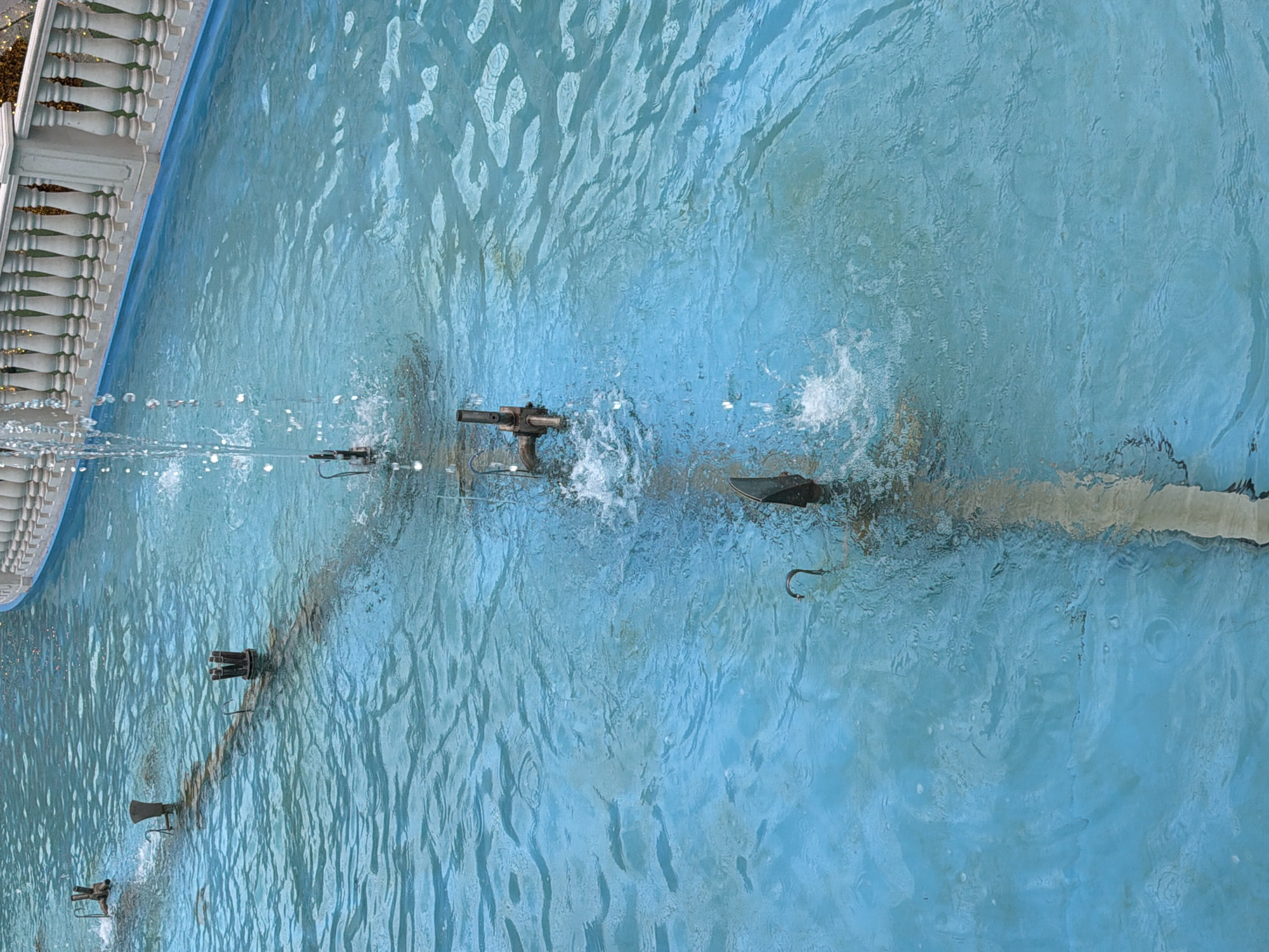 An aerial view of a shallow, rectangular fountain in an urban environment shows a row of metal spouts embedded in the blue water, intermittently spraying water. The fountain's edge is lined with a white balustrade, characteristic of decorative outdoor spaces. The water is clear, revealing the dark bottom of the fountain and the metal pipes.