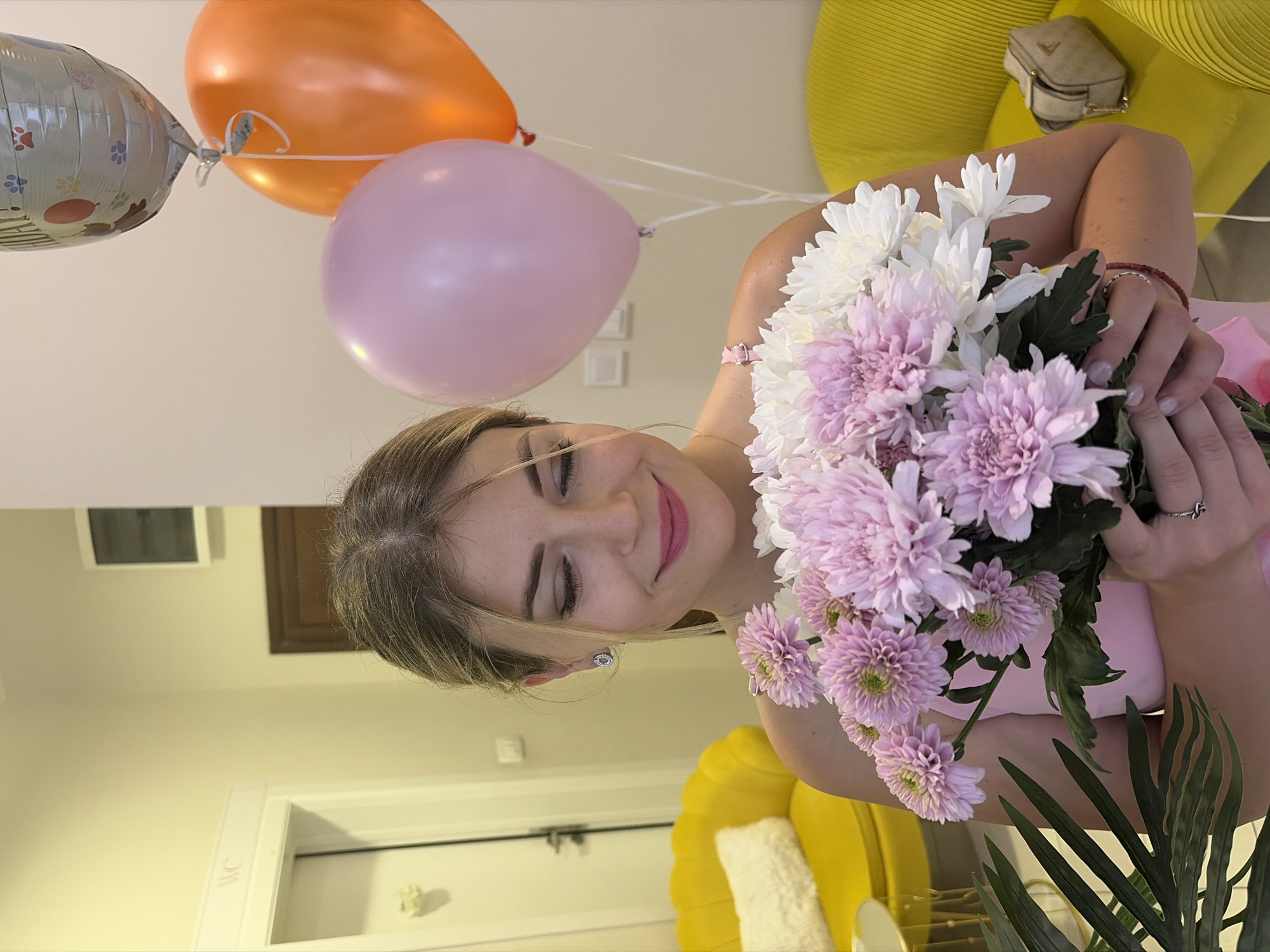 A young woman with blonde hair pulled back is holding a bouquet of pink and white chrysanthemums. She is wearing a pink dress and a silver earring in her left ear. Her eyes are closed, and she has a gentle smile. In the background, three balloons are visible: a silver one with a "Happy Birthday!" message and paw prints, a pink one, and an orange one. To the left of the woman, a bright yellow armchair with a white pillow is partially visible, and to her right, another yellow armchair with a small beige purse on it. The setting appears to be an indoor space, possibly a home or hotel room, with light-colored walls and a doorway visible on the left. The overall impression is one of a celebratory occasion.