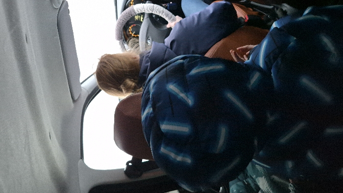 Inside a vehicle in the city of Ol'khovochka, Russia, a view is presented from a rear passenger seat looking forward. In the immediate foreground, the back of a person, likely a child, is visible, wearing a dark blue puffer jacket with irregular light blue stripe patterns. In the midground, the back of an adult is seen in the driver's seat, with light brown hair pulled into a bun. This individual wears a dark blue jacket, and their right hand is visible on a steering wheel fitted with a grey, fluffy cover. The vehicle's dashboard is partially visible, displaying an illuminated instrument cluster with orange indicators. Beyond the windshield, the exterior environment appears uniformly bright white, suggesting overcast or snowy conditions. The vehicle's interior ceiling is light grey fabric, and a passenger-side sun visor is retracted.
