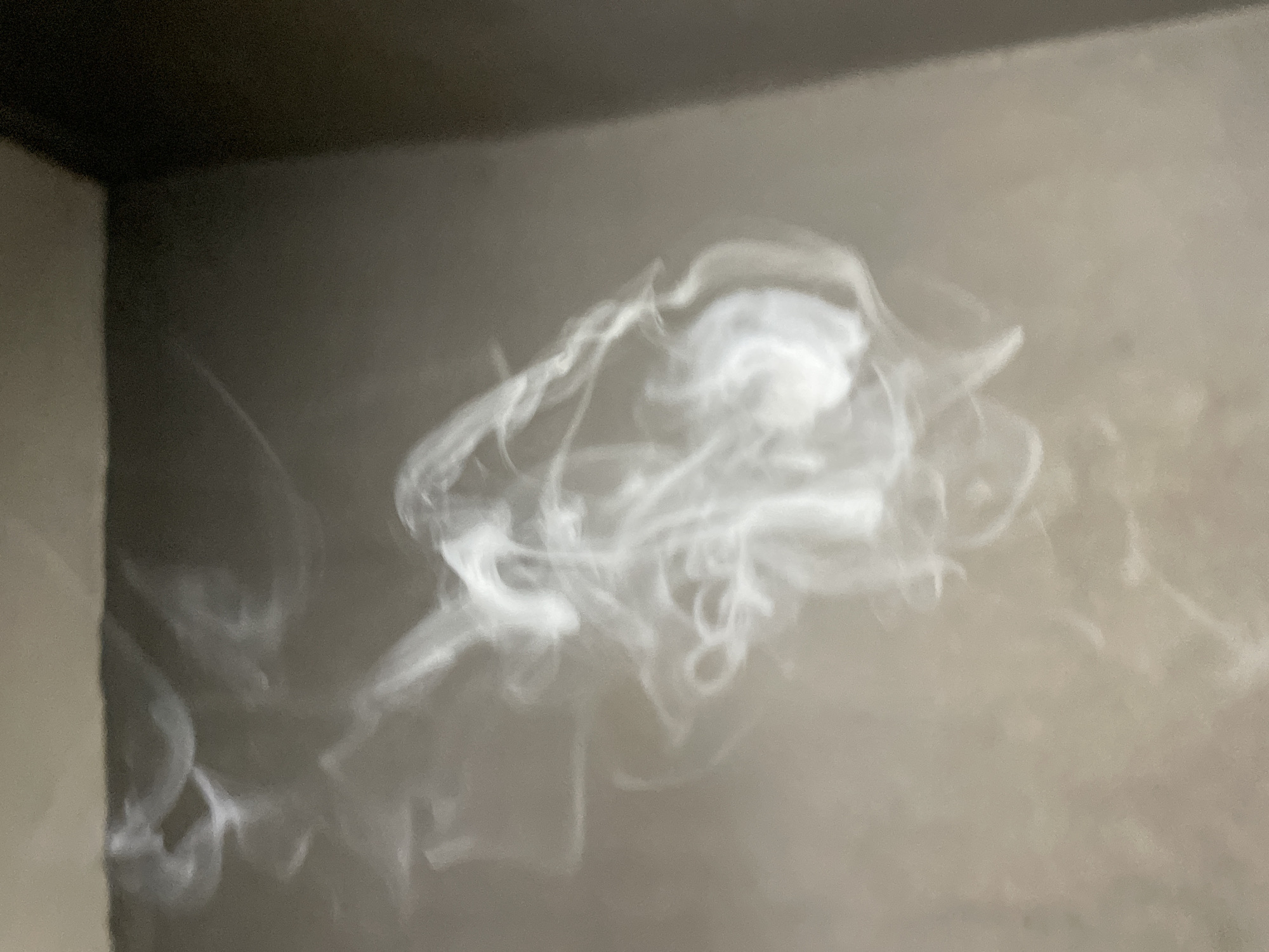 The image is a close-up, slightly blurred photograph of white smoke swirling against a muted, textured background. The smoke forms amorphous, wispy shapes, with some areas denser and brighter than others. The background appears to be a neutral-colored surface, possibly a wall or a countertop, with a darker vertical element visible on the right side, suggesting a corner or edge. The lighting is diffused, casting soft shadows. There are no visible people, specific objects, or discernible actions in the scene, other than the movement of the smoke itself. The image does not contain any information that would allow for identification of a specific location within Nkpor, Nigeria, or any other geographic area.