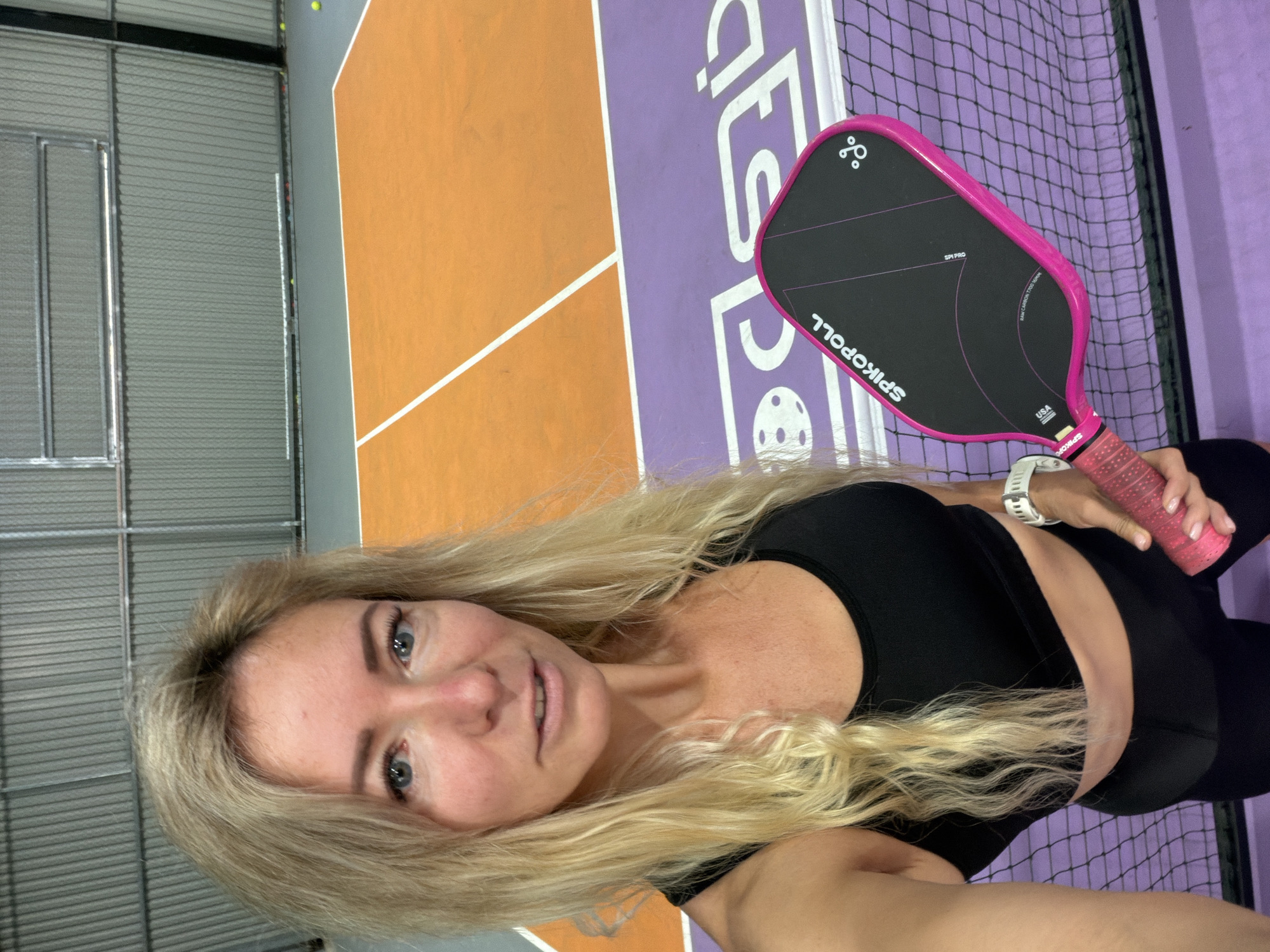 A woman with long blonde hair, wearing a black sports bra and black leggings, stands on a pickleball court. She is holding a pink pickleball paddle in her right hand and has a white watch on her left wrist. The court has orange and purple sections with white lines. In the background, there is a grey corrugated metal wall and a fence. The scene appears to be indoors, possibly in an athletic facility.