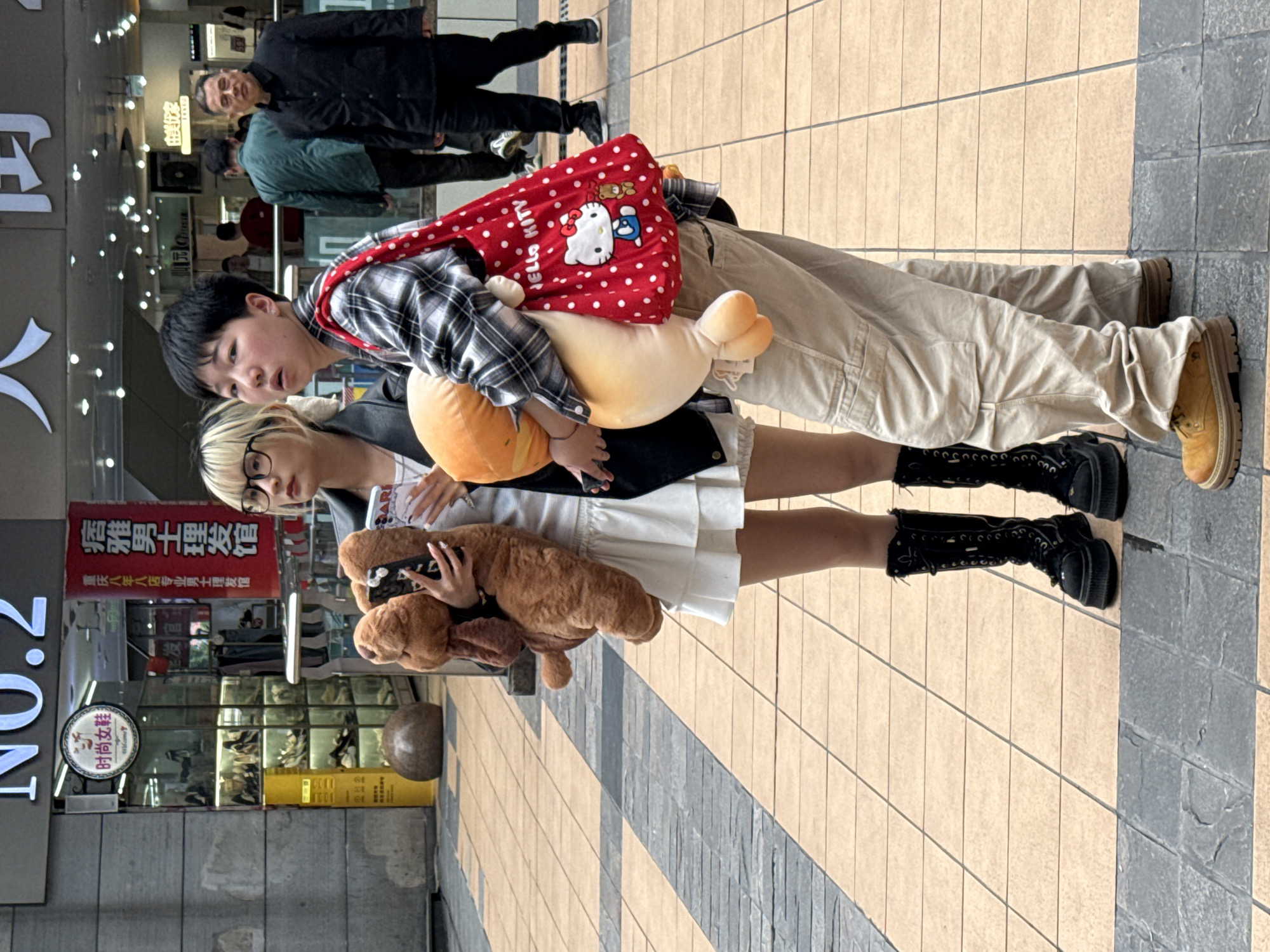 Here is a concise, forensic description of the scene:
Two people stand on a paved city street in Liangjiang, China, posing for a photograph. The person on the right is wearing beige cargo pants, a plaid shirt, and yellow boots. They are carrying a red Hello Kitty bag and an orange, oblong plush toy. The person on the left is wearing a white dress, a black jacket, black boots, and glasses. They are holding a large brown stuffed dog. A shoe store can be seen in the background, along with a sign for a barber shop. Pedestrians are visible further in the background.