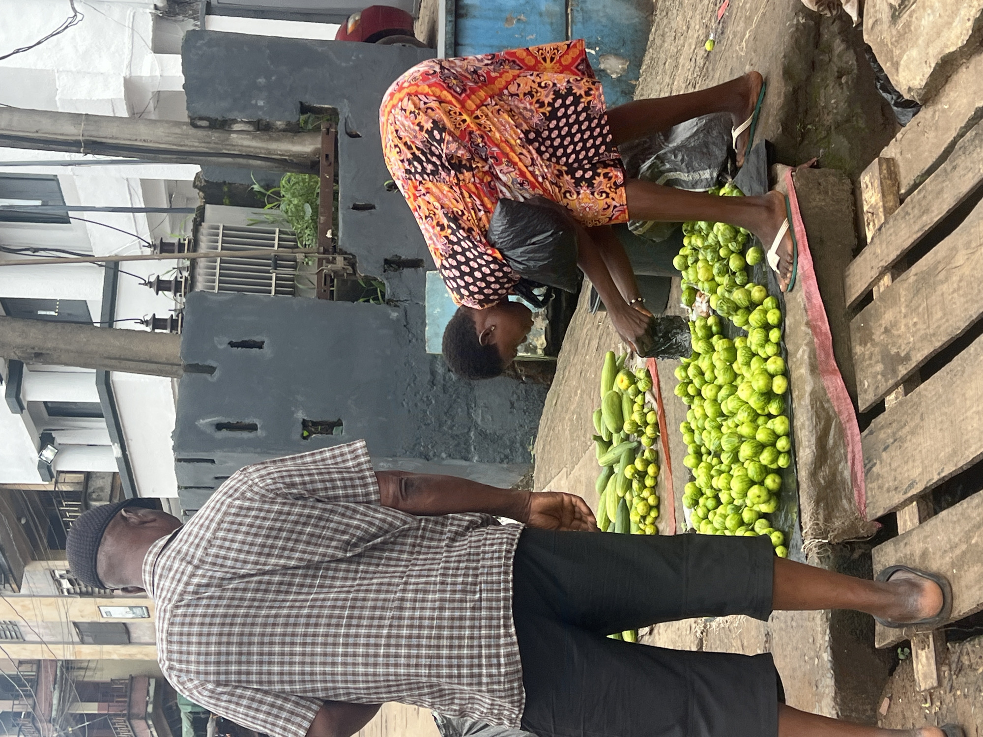 In Nkpor, Nigeria, a man wearing a checkered shirt and black shorts stands with his back to the camera, facing a woman in a colorful dress who is bent over a large pile of green vegetables. The woman is holding a black plastic bag in her left hand and is reaching for more produce. The vegetables, appearing to be cucumbers and possibly small gourds, are spread across a red plastic sheet on the ground. In the background, a building with multiple stories and windows is visible, along with electrical wires and a transformer. The scene is set on a rough, uneven surface, with wooden planks placed in the foreground. A red car is partially visible on the right side of the image.