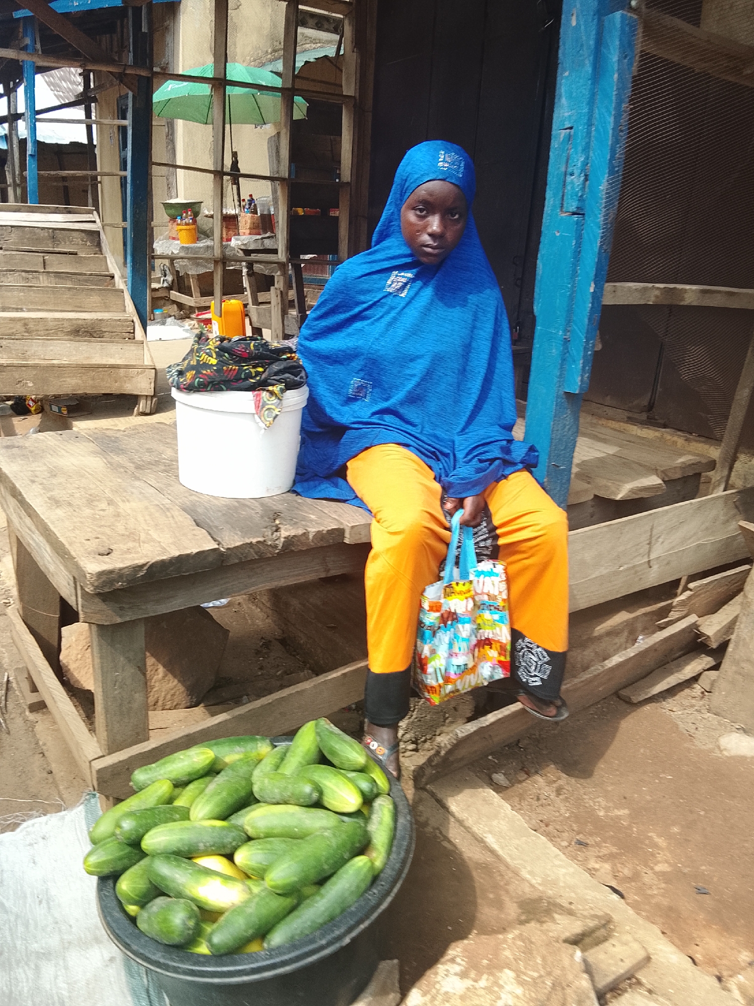 In Akwanga, Nigeria, a young woman wearing an azure hijab and bright orange trousers sits on a rustic wooden bench outside a market stall. She holds a colorful patterned shopping bag in her right hand. A white bucket containing fabric is placed on the bench beside her. In front of her, a large black tub overflows with freshly harvested cucumbers. The background reveals a marketplace setting with wooden structures, stairs, and stalls, suggesting a busy commercial area.