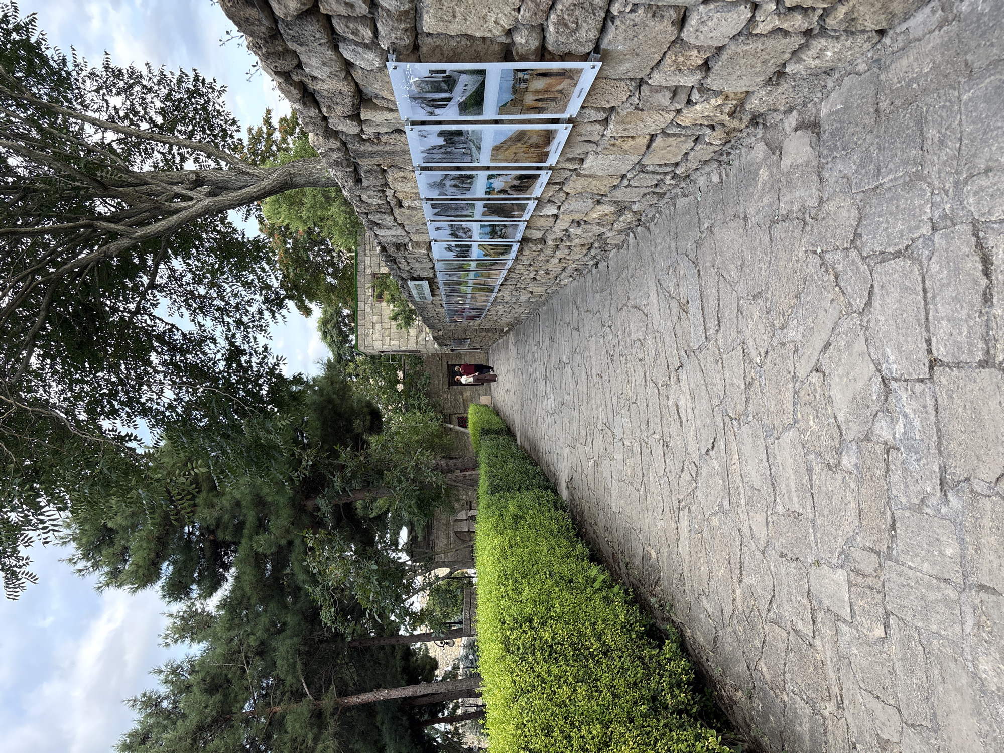 The media file depicts an outdoor scene in Derbent, Russia, featuring a stone wall with framed photographs attached to it. The photographs are arranged in a horizontal line, with multiple images visible. The wall is constructed of rough, grey stones, and a paved walkway runs alongside it. To the left of the walkway, a meticulously trimmed green hedge lines the path. Beyond the hedge, trees with dense foliage and a clear sky with scattered clouds can be seen.
In the distance, along the walkway, two individuals are walking away from the camera. They appear to be casually dressed. The scene suggests a public space, possibly a park or a historical site, where an exhibition of photographs is being held. The lighting indicates it is daytime, likely late morning or early afternoon, with bright but diffused light, suggesting overcast or partly cloudy conditions. The overall atmosphere appears peaceful and contemplative, with the natural elements of trees and greenery contrasting with the man-made structures of the wall and walkway. No visible text is discernible in the media file, except for potentially within the photographs themselves, which are too small and distant to read.