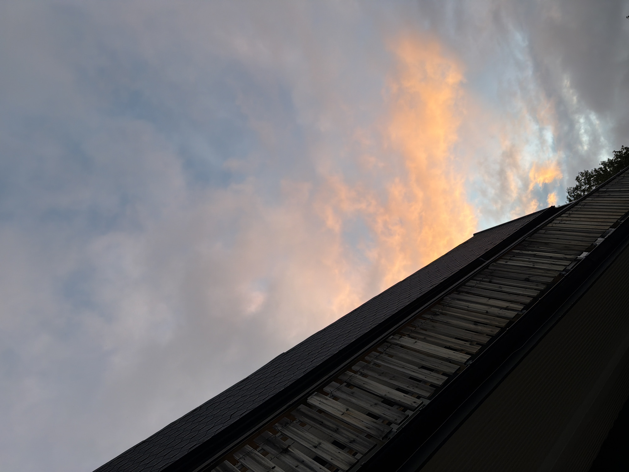 The image captures a low-angle view of a building's roof and a wooden railing against a sky filled with clouds. The roof is dark and appears to be shingled. The railing, made of vertical wooden planks, is in shadow, showing some texture and wear. In the sky, clouds are illuminated with soft, warm hues of orange and pink, suggesting a sunset or sunrise. The background sky transitions to cooler tones of blue and gray. Some green foliage from trees is visible at the bottom right of the frame, partially obscured by the building. This scene is likely a residential or commercial building in Harstad, Norway, given the context. No people or specific actions are depicted.