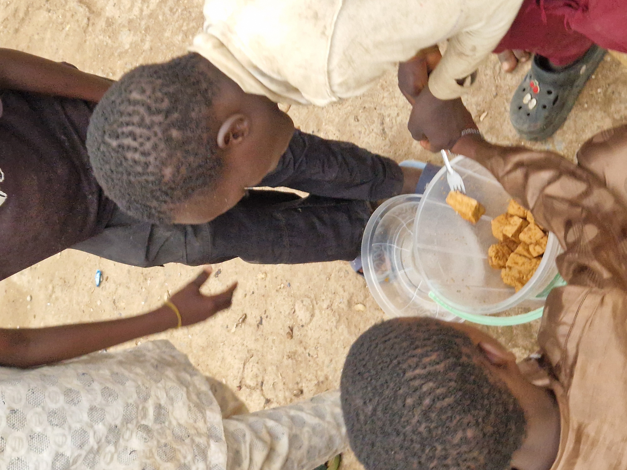 In Jos, Nigeria, three young individuals are gathered around a clear plastic container filled with cubed food. One person, wearing a white garment with a patterned design, stands on the left. Their arm, adorned with a yellow bracelet, is extended towards the container. To their right, another person in dark clothing leans over, their head partially visible. A third individual, in a beige garment, is on the right, actively engaging with the food. They are holding a fork, which has a cube of food impaled on it, and their hands are reaching towards the container, possibly to take more food. The ground is sandy and appears to be an outdoor setting, with small debris scattered around. One person is wearing a dark croc-style sandal with colorful charms. The overall scene suggests a moment of shared meal or snack in an informal, likely outdoor, environment.