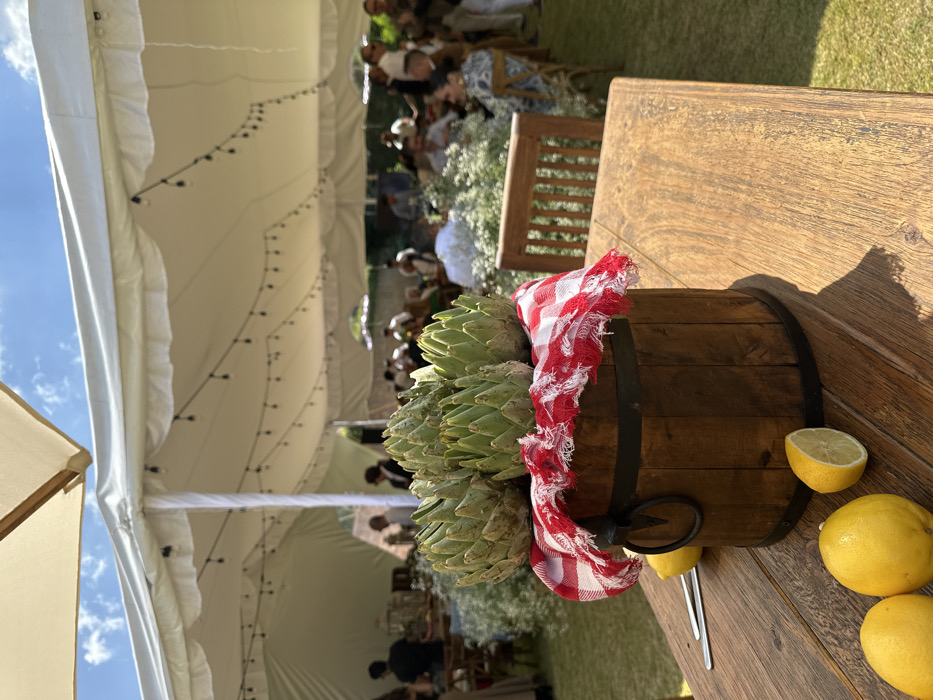An outdoor event is depicted under a large white tent on a sunny day in the city of Unknown, Unknown. In the immediate foreground, a rustic wooden table holds a centerpiece: a dark brown wooden barrel with two black metal bands and a single black metal ring handle. Draped over the barrel's opening is a red and white gingham-patterned cloth with frayed edges, containing five whole green artichokes. To the left on the table are three whole yellow lemons and one yellow lemon cut in half, exposing its flesh. Two silver utensils are visible beside the lemons.
The midground features a green lawn area populated with numerous people, approximately two dozen, seated and standing at various wooden tables under the tent. White floral arrangements are visible on some tables. Individuals appear to be socializing or dining; their faces are largely indistinct. A slatted wooden chair back is partially visible on the right side of the frame.
The background is dominated by the expansive white canvas tent, supported by poles. String lights with exposed bulbs hang in rows across the underside of the tent's ceiling. Beyond the tent's perimeter, a bright blue sky with scattered white clouds is visible, along with blurred green foliage, indicating an open-air setting. Sunlight casts defined shadows from the foreground objects onto the wooden table.