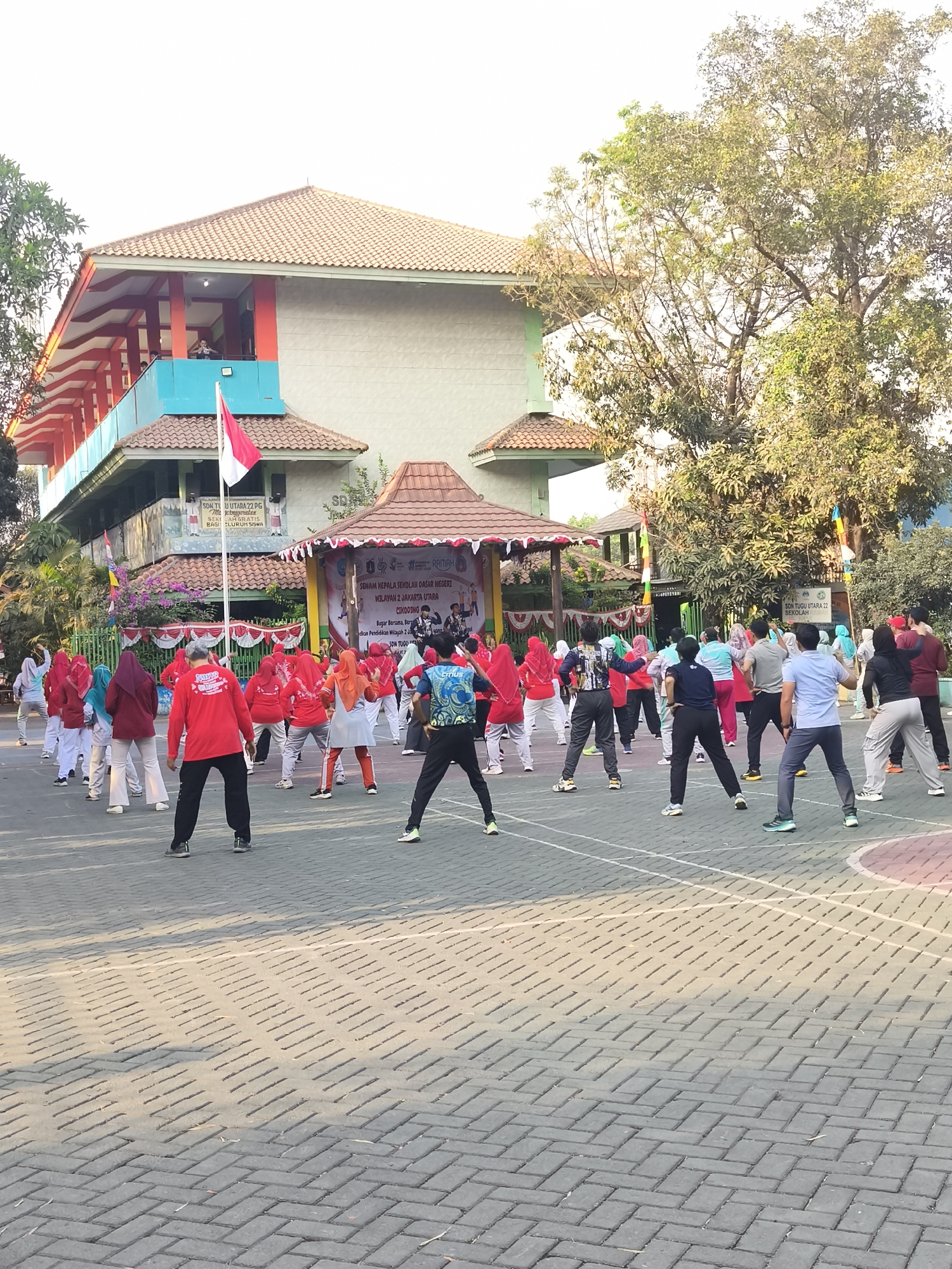 In North Jakarta, Indonesia, on a paved outdoor court, a group of people are engaged in an exercise activity. The scene is set in front of a two-story school building with a red and white striped awning and a red tiled roof. A prominent Indonesian flag is visible flying from a pole in front of the building. Several individuals are dressed in bright red shirts and white pants, participating in synchronized movements. Other participants are wearing a variety of colored athletic wear. The background features trees and a banner with Indonesian text, suggesting an organized event or class. The time of day appears to be morning or late afternoon, indicated by the lighting and the long shadows cast on the court.