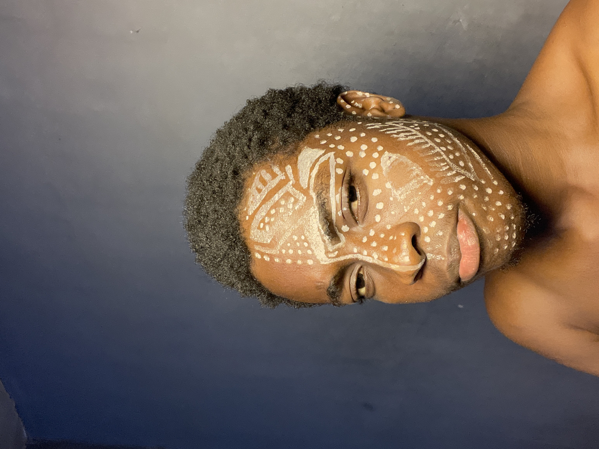 A close-up portrait of a Black male with short, dark, curly hair. His skin is adorned with white painted patterns, including dots and linear designs, possibly tribal or ceremonial markings. He is looking slightly down and to the right. The background is a blurred, dark gradient, suggestive of a wall or backdrop. There is no information regarding the location of this picture within Nkpor, Nigeria.