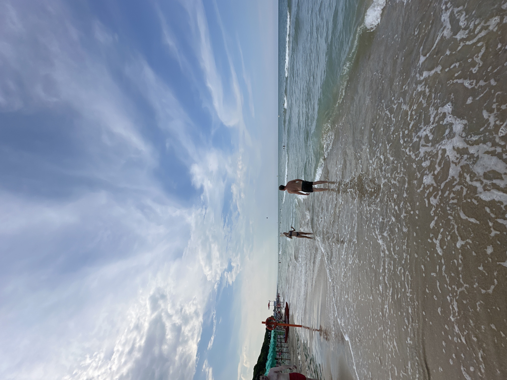 The image captures a beach scene in Pineto, Italy, likely during the daytime given the bright sky and visible sunlight. The perspective is from low down on the sand, looking towards the sea and sky, with the image rotated such that the horizon is vertical.
The foreground shows the wet sand with incoming waves creating frothy patterns. Two people are visible near the waterline. Further out in the sea, several more people can be seen, some wading and others further offshore. The sea itself appears calm with gentle waves.
The sky is a prominent feature, dominated by a vast expanse of blue with wispy, elongated white clouds that stretch across the frame. These clouds suggest a partly cloudy but pleasant day, possibly with a light breeze.
In the middle ground, closer to the shore, there are signs of a beach setup. A red and orange life raft with a lifebuoy attached is visible, supported by a stand, indicating a lifeguarded area. Beyond this, beach umbrellas and some vegetation can be seen, suggesting a developed beach with amenities.
The overall mood of the image is relaxed and summery, capturing a typical day at the beach with people enjoying the water and the coastal environment. There is no visible text in the image.