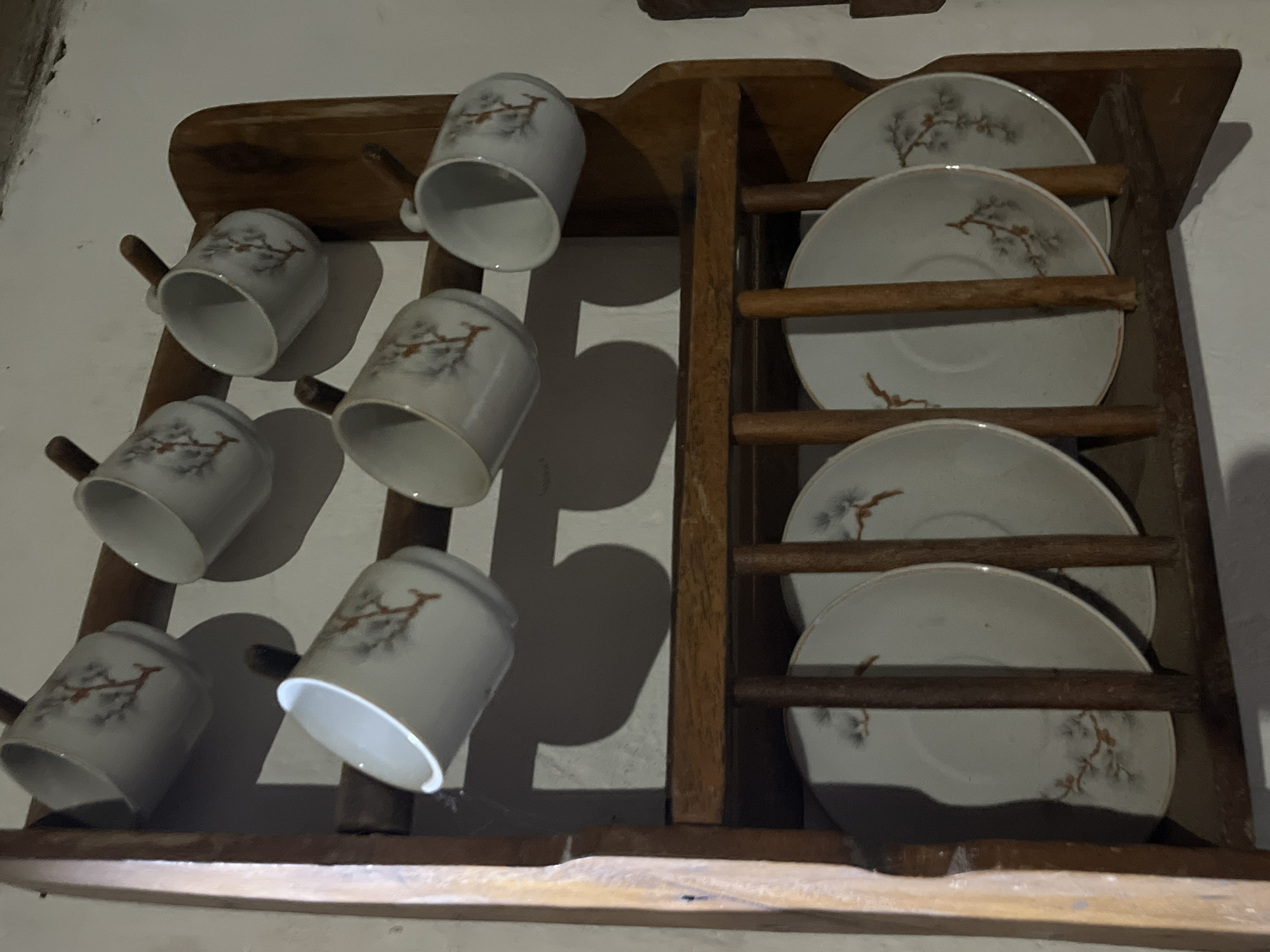 This is a close-up, eye-level shot of a wooden wall-mounted dish rack holding six white teacups and five saucers. The teacups are adorned with a delicate pattern of brown branches with small gray leaves. The saucers, also white, have a similar branch design. The dish rack is composed of dark wood, with a side panel visible on the left, and two horizontal shelves for the cups and saucers. The background is a neutral, light-colored wall. The scene appears to be an interior domestic setting. This image does not provide enough information to determine the location in Juan Griego, Venezuela.