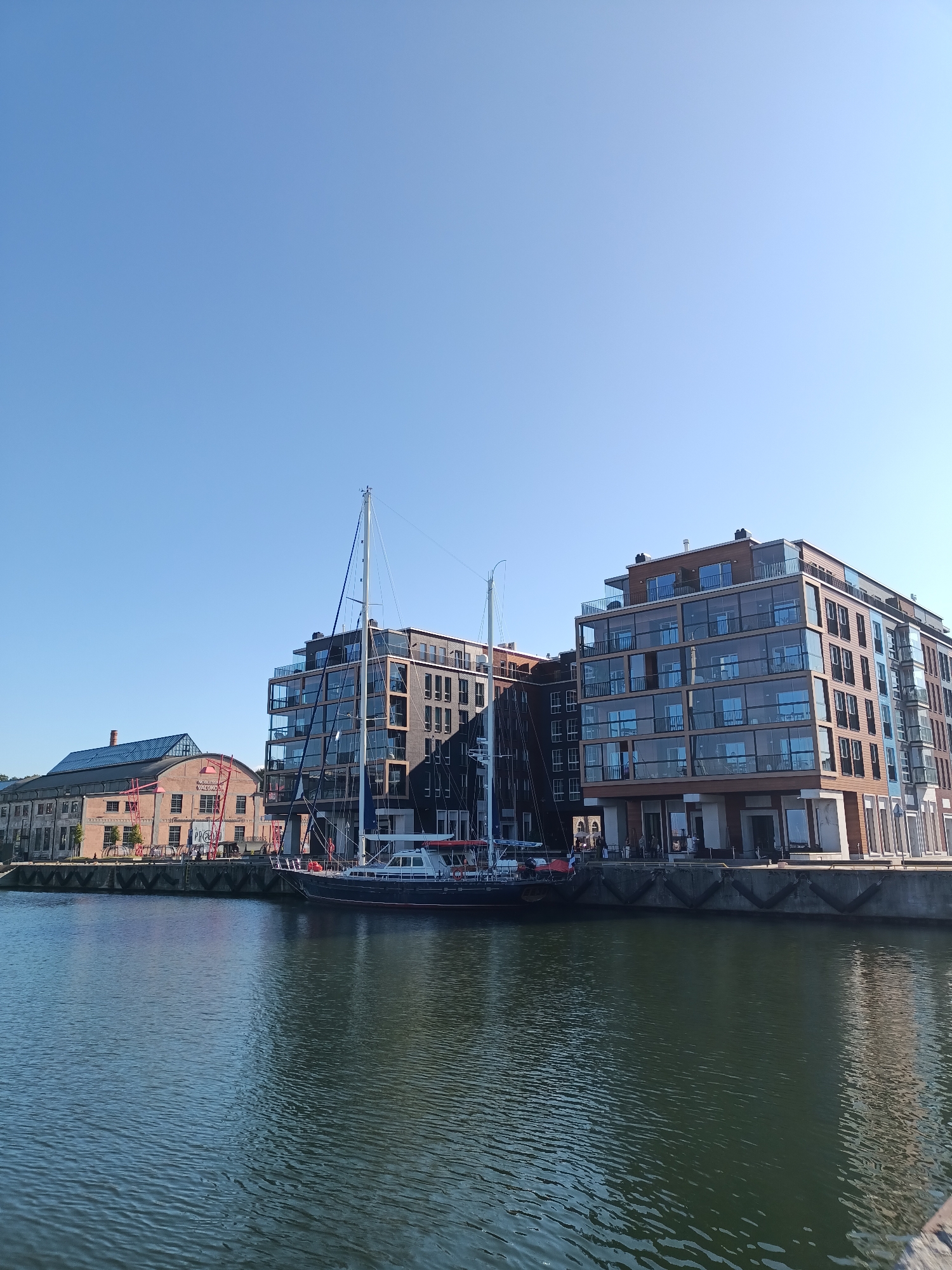 The image depicts a waterfront scene in Tallinn, Estonia, likely the Old City Harbor area, on a clear, sunny day. In the foreground, a calm body of dark green water reflects the surrounding structures and the sky. Moored along the quay is a dark-hulled sailing yacht with tall masts.
Behind the yacht, a row of modern, multi-story buildings with extensive glass facades and wooden accents lines the waterfront. These buildings have balconies and multiple windows, suggesting residential or office spaces. To the left of these modern structures, an older, brick building with a distinctive blue roof and industrial-style windows is visible. A red industrial crane or structure with the word "PRO" visible on it is also present near the older building.
Several people can be seen walking along the quay in front of the buildings, enjoying the waterfront. The sky is a clear, bright blue with no clouds. The overall impression is one of urban development and modern architecture integrated with historic elements in a harbor setting.