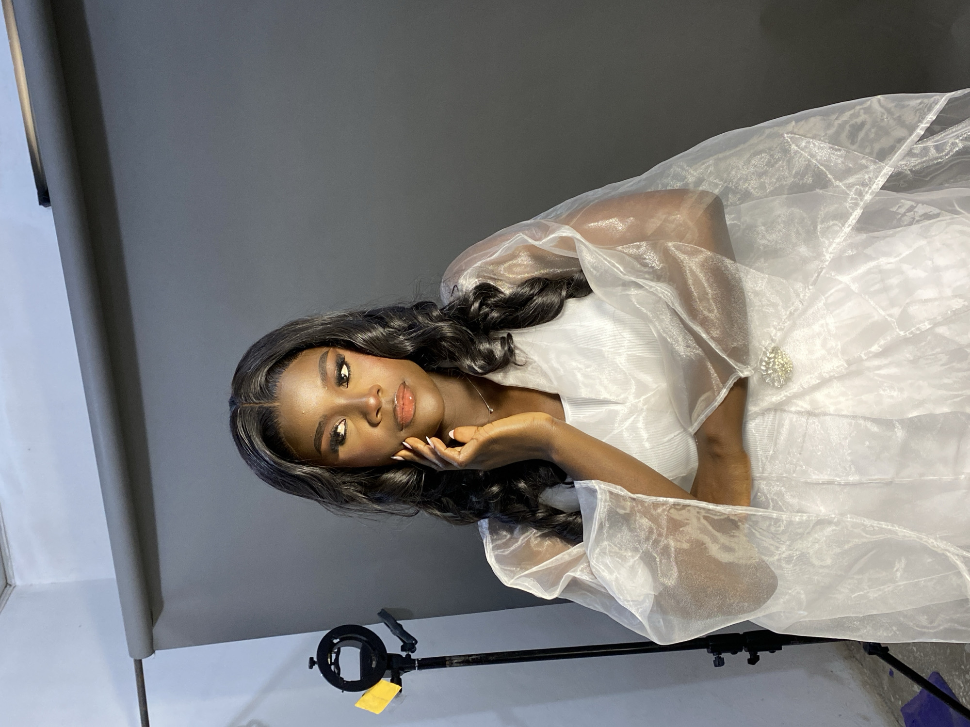 A young woman with dark skin and long, dark wavy hair poses in a white dress with a sheer overlay. Her face is close to the camera, showing her makeup with glossy lips and dark eyeliner. Her hand is raised near her face. The backdrop is a simple grey surface. A lighting stand is visible in the lower part of the frame. The location appears to be indoors in a studio setting and there is no information available to identify the city.
