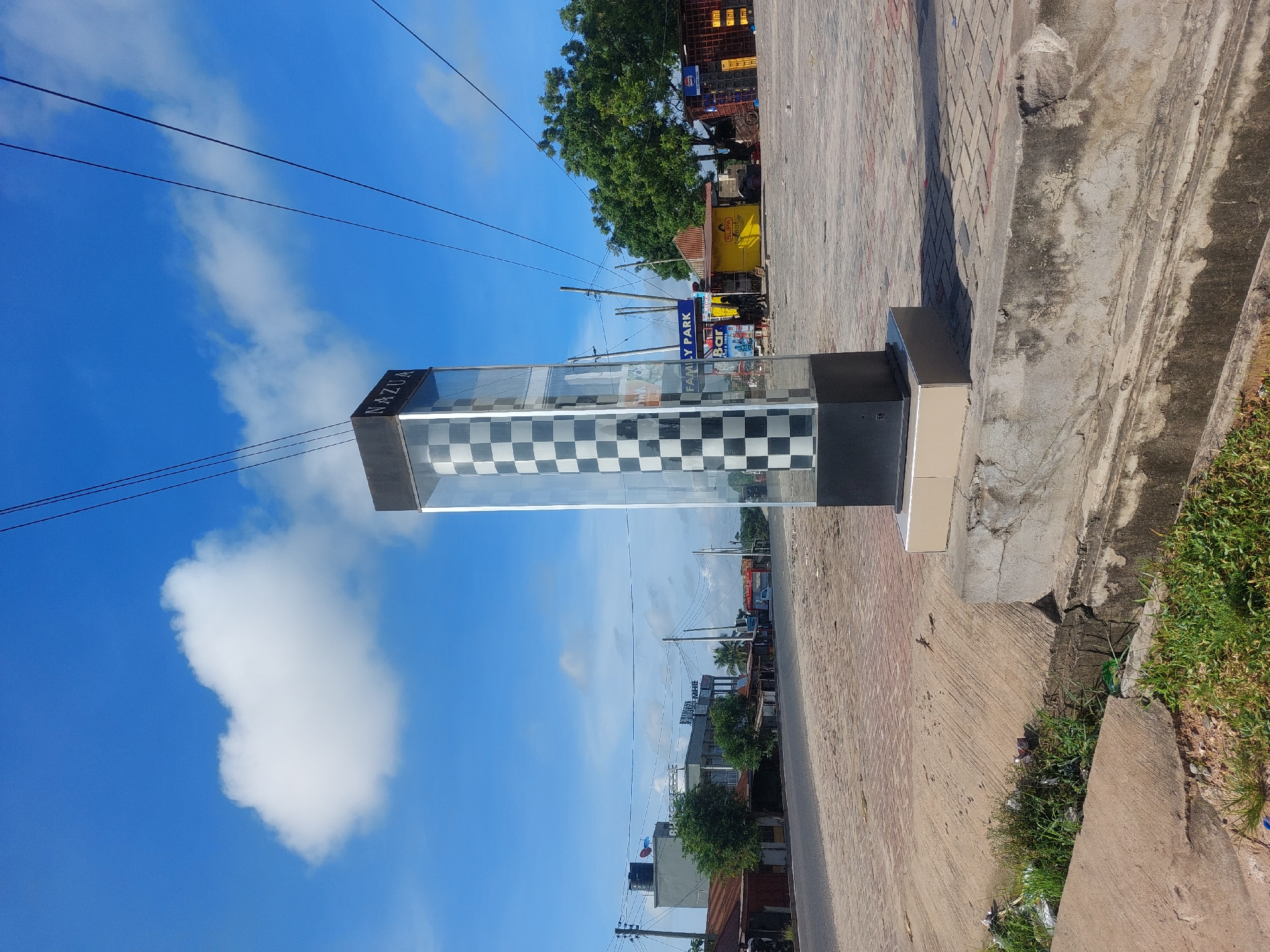 The image shows a street scene in Dar es-Salaam, Tanzania. A tall, rectangular, glass display case with a black and white checkered pattern is situated on the edge of a sidewalk. The top of the display case is labeled "NAZUA." The sky is blue with scattered white clouds. Power lines crisscross the sky. The sidewalk is made of interlocking pavers and abuts a concrete curb with vegetation growing along the edge. Across the street, buildings and trees are visible, including storefronts with signage in the local language. The street appears relatively empty of traffic.