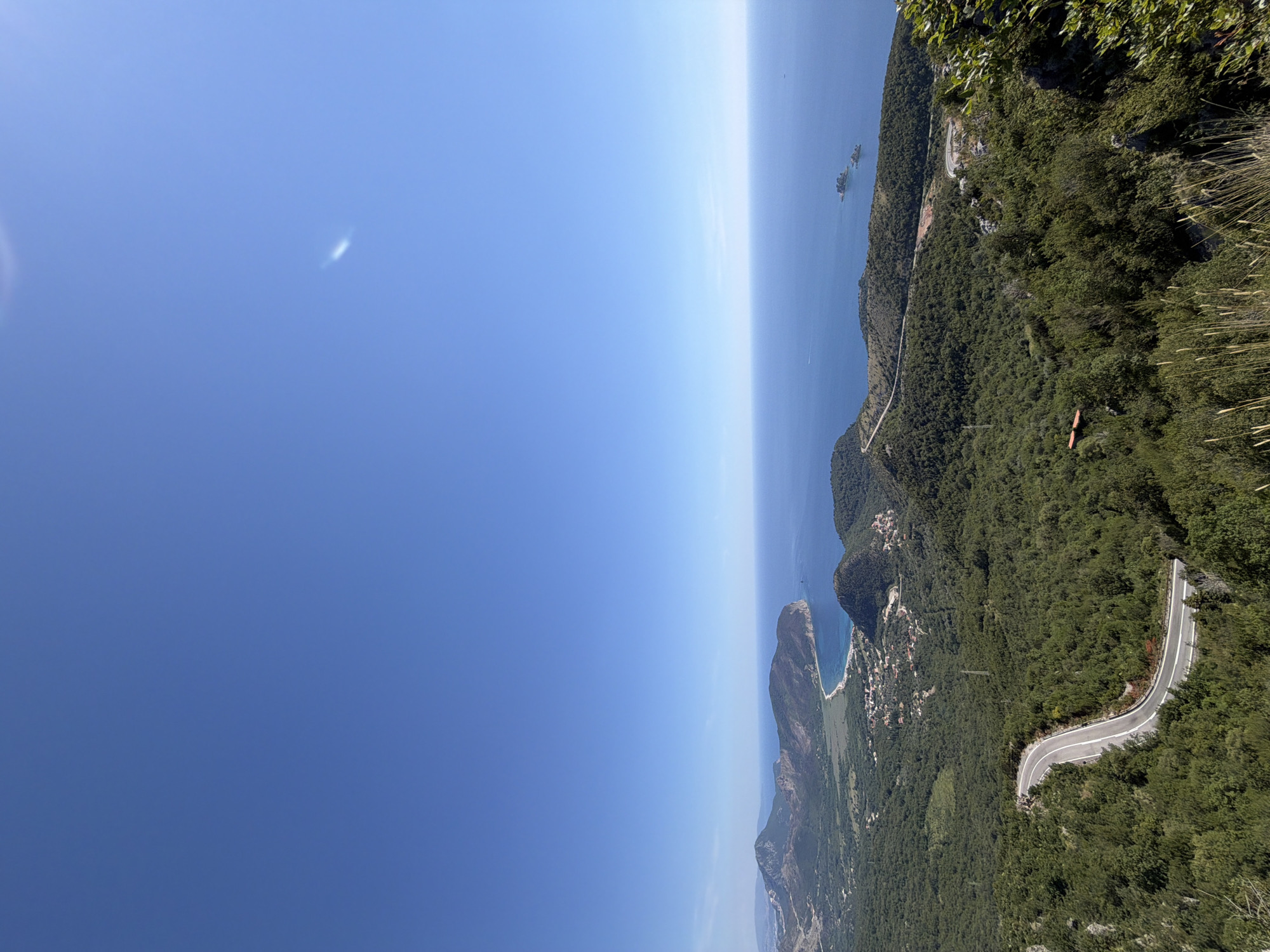 The image depicts a panoramic view of a coastal mountainous region under a clear blue sky with a wispy cloud formation. The foreground is dominated by a winding road cutting through dense green foliage. The road features white lane markings and appears to be a paved asphalt surface. In the mid-ground, a valley opens up, revealing a small settlement with red-tiled roofs nestled amongst the green landscape. Further in the distance, the coastline meets the deep blue sea, with a curved beach visible. Several peninsulas and hilly terrains extend into the sea. Two small islands are situated offshore, with a boat visible in the water near them. The overall scene suggests a natural landscape with a human presence in the form of a road and a small village. The location is identified as Novoselje, Montenegro.