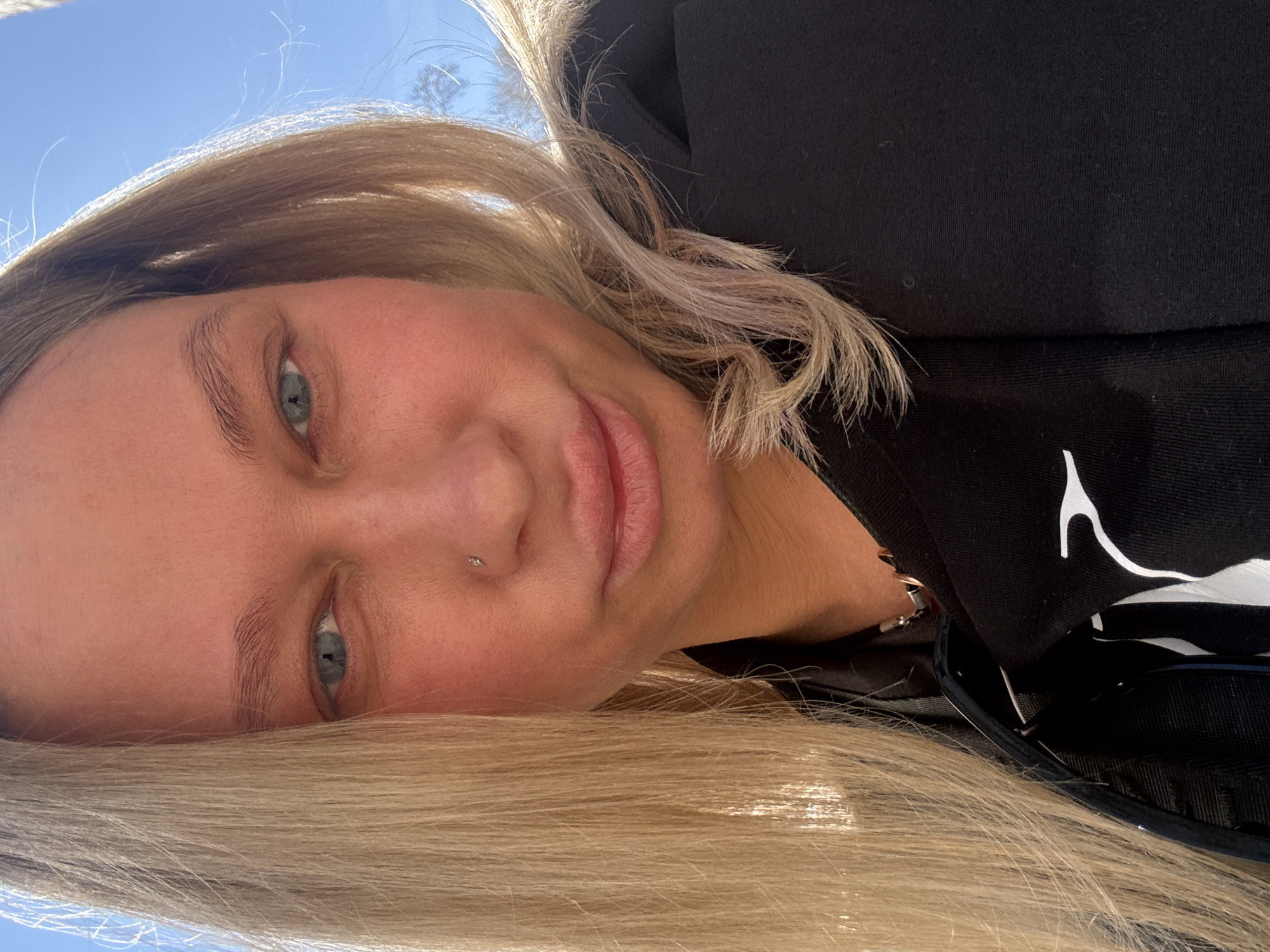 A selfie image of a Caucasian woman with blond hair and fair skin. She has blue eyes and a small stud piercing on the left side of her nose. She wears a black top with a white graphic design on the upper right. The photo appears to be taken outdoors under a blue sky. Location not discernible from image.