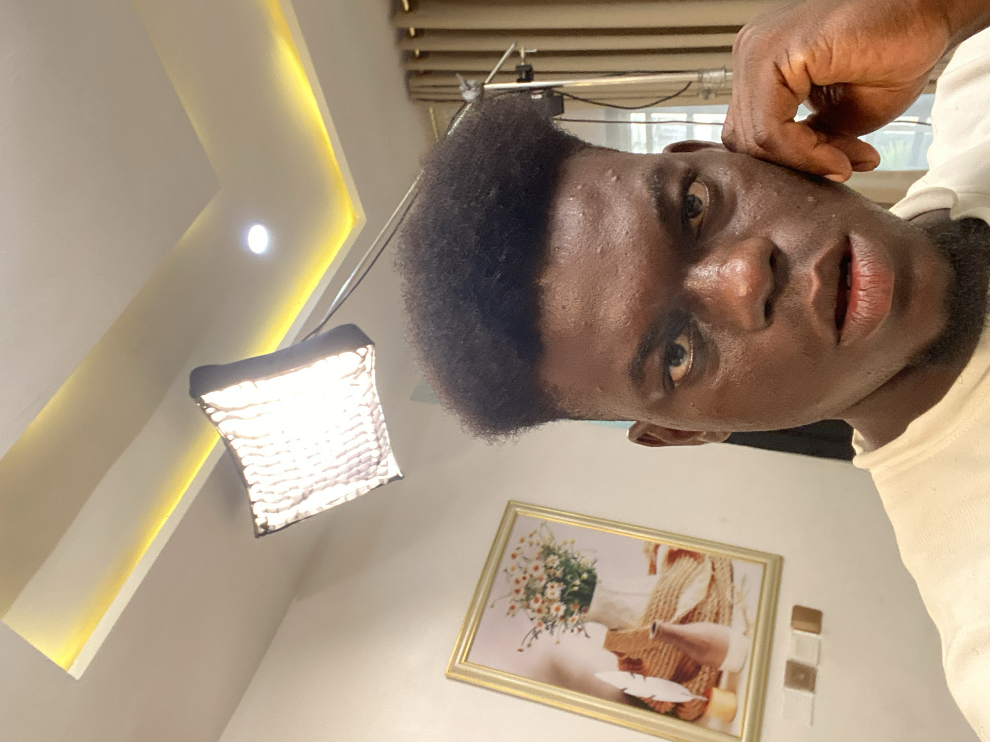 A low-angle shot captures a man's face, slightly angled to the right, with his hand touching his ear. The man has dark skin and closely cropped black hair. His expression appears somewhat intense. In the background, a photographic light is visible on the left, casting a bright glow. Behind the man, a framed picture hangs on the wall. The location appears to be indoors, characterized by a neutral-colored wall and ceiling with decorative lighting.
