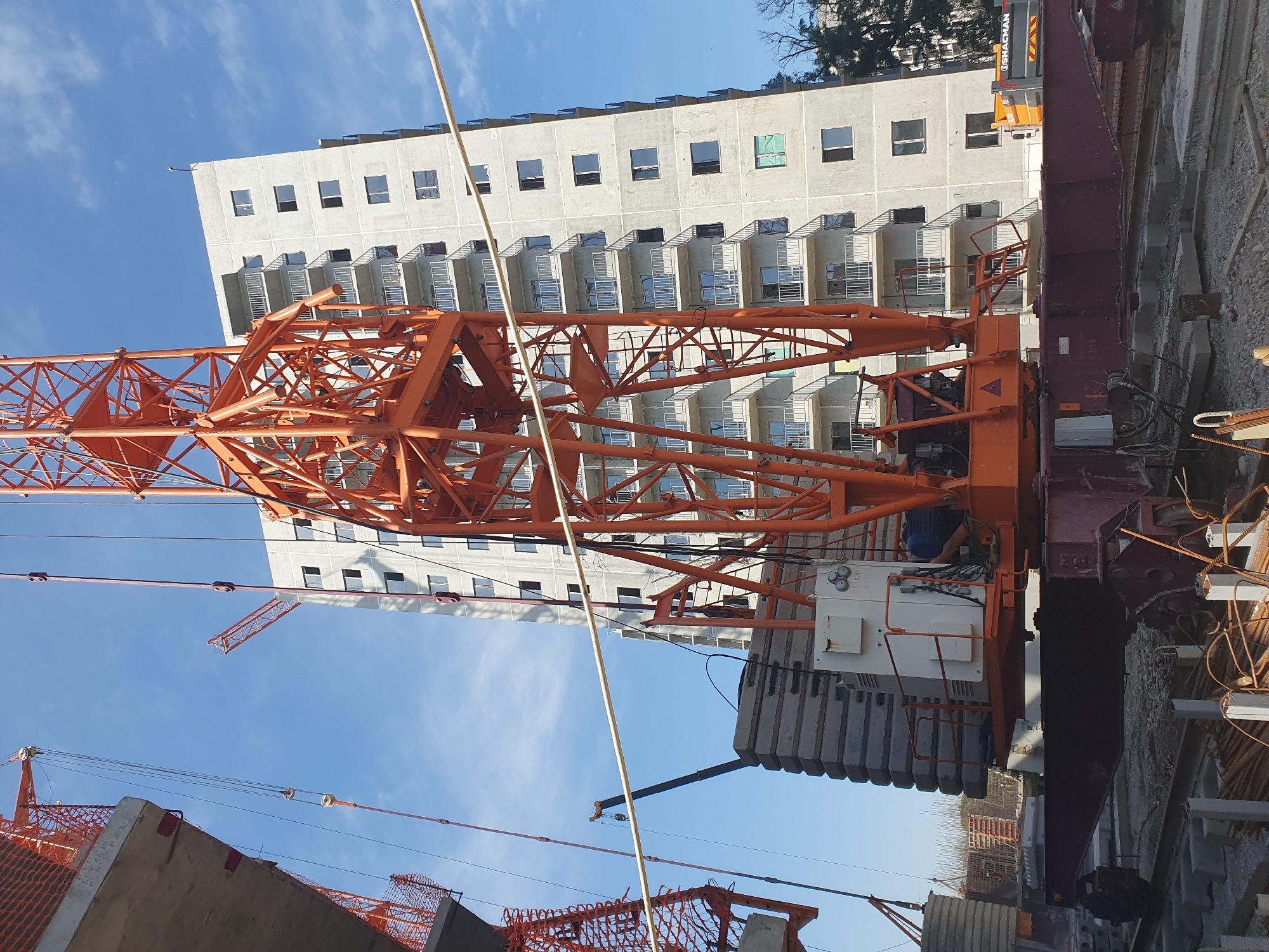 An external, eye-level, medium shot captures a construction site under a bright blue sky with scattered clouds, featuring an unfinished apartment building and construction equipment in Tashkent, Uzbekistan. In the foreground, a large orange crane dominates the frame, its intricate metal latticework visible against the sky. The crane's base rests on a dark red chassis with visible wheels and tracks, and a stack of large, rectangular concrete slabs is positioned behind it. A white control box is mounted on the crane's base. To the left and partially obscured, another construction crane is visible, also with orange elements and netting. Further left, a concrete structure, possibly a floor or ceiling slab of another building, juts into the frame.
Behind the crane, the tall, grey concrete apartment building rises, its facade marked by rows of windows and balconies with metal railings. The building appears to be in an advanced stage of construction, with some windows already installed. A smaller orange construction crane can be seen in the distance, extending above the apartment building. To the far right, a portion of a truck with the branding "SHACMAN" is visible, suggesting ongoing logistical operations. The ground is covered with construction debris, including gravel, concrete pieces, and rebar. The overall scene depicts an active construction environment in a city setting.