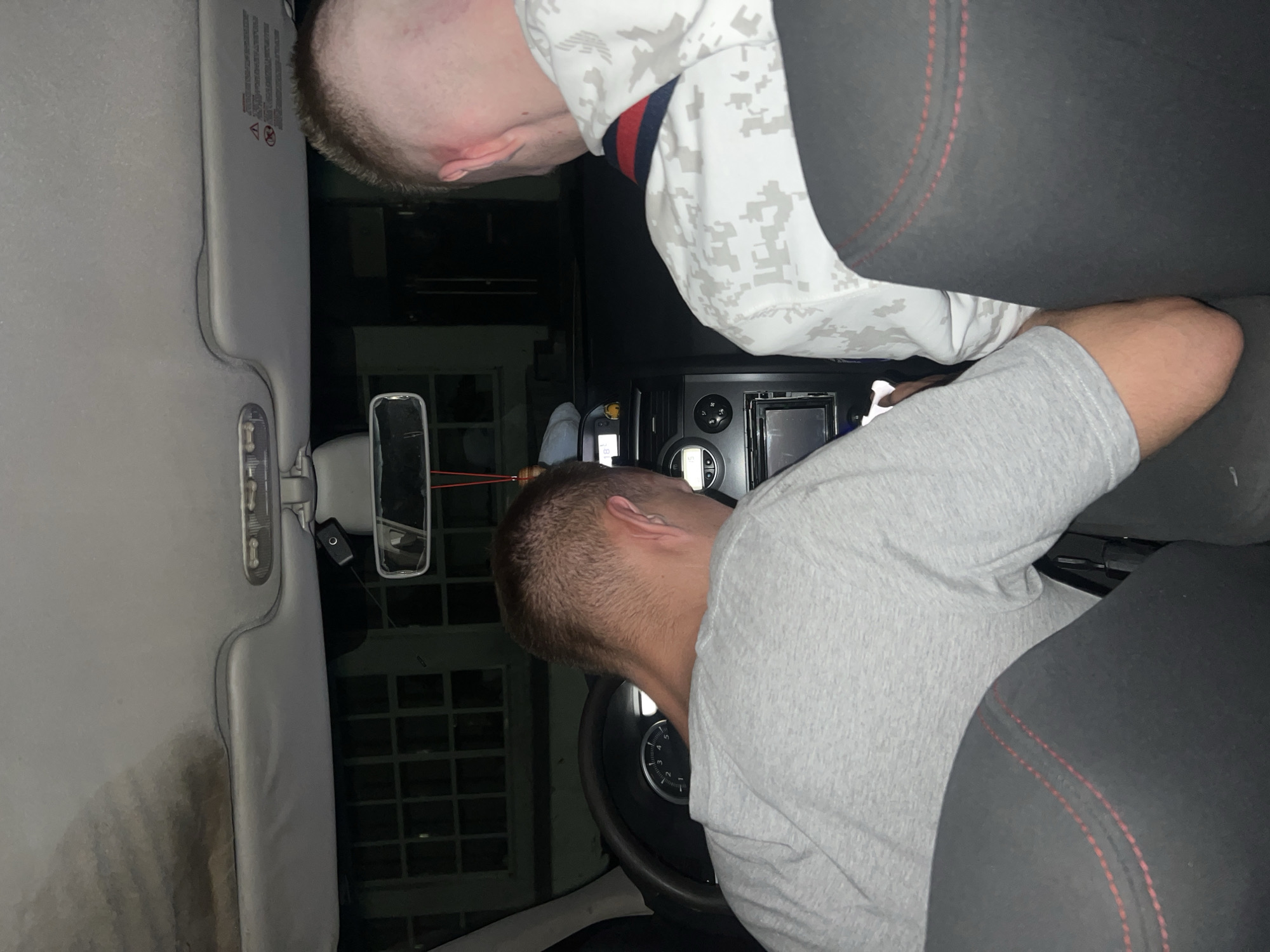 The scene is set inside the front cabin of a vehicle at nighttime in the city of Ветлужский, Russia, viewed from the rear passenger side. Two adult males occupy the front seats. The male in the driver's seat, visible from the back of his head and neck, has short, light-brown hair and wears a light grey short-sleeved t-shirt. He is leaning forward, directing his gaze downward towards the center console area, with his right hand appearing to interact with a device. The male in the front passenger seat, also visible from the back of his head and neck, has short, light-brown hair and wears a white hooded sweatshirt with a grey digital camouflage pattern and a horizontal red and navy blue stripe across the upper back.
The car's interior features include a significantly stained and discolored headliner above the driver's seat. An overhead console with two transparent light covers and sun visors, one displaying warning labels, are present. A small, black electronic device, possibly a dashcam, is suspended from the rearview mirror by a red cord. The dark-colored steering wheel is partially visible, along with an instrument cluster showing RPM markings. The center console contains an infotainment screen, control buttons, air vents, and a small digital display showing "18°C". The front seats are dark-colored with red stitching visible on their contours. Outside the vehicle, a building with multi-pane windows is dimly visible through the windshield and side windows, suggesting an urban or suburban environment.