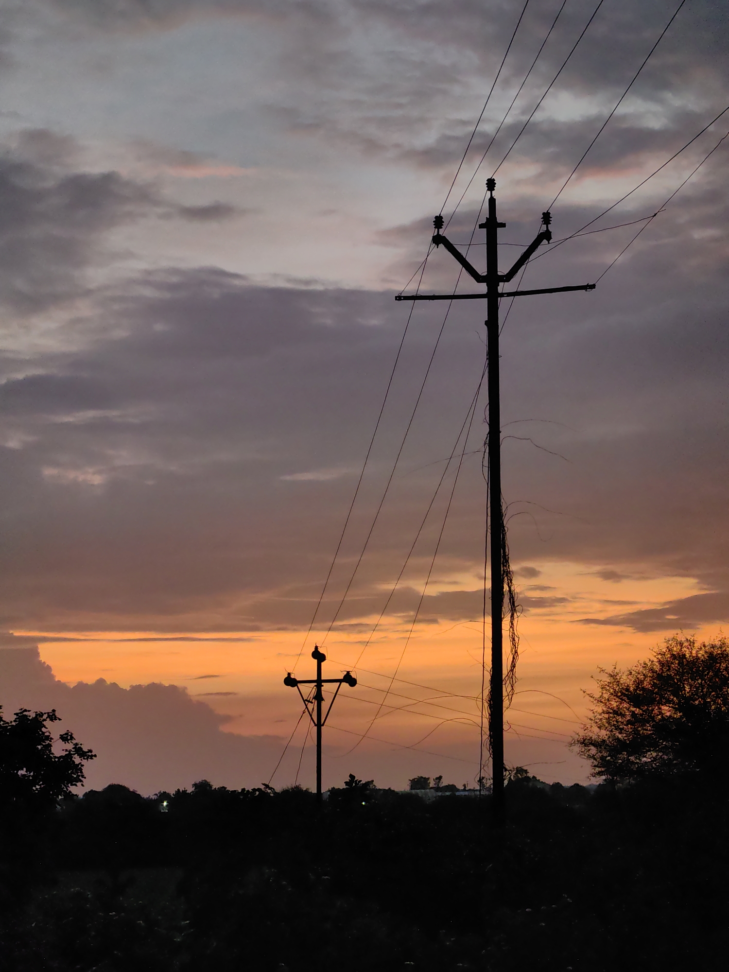 This is a silhouetted image of a sunset in Dattapur, India, with two utility poles and power lines prominently featured against a dramatic sky. The sky displays a gradient of colors, transitioning from a vibrant orange and yellow near the horizon to muted purples and grays higher up, with scattered clouds. The foreground is dark and obscured, showing the silhouettes of trees and vegetation. The utility poles are tall and black, with wires extending between them and across the frame. One pole is closer and larger, while another is smaller and further in the distance. There are no people or animals visible in the scene. The overall impression is one of a rural or semi-rural landscape at dusk.