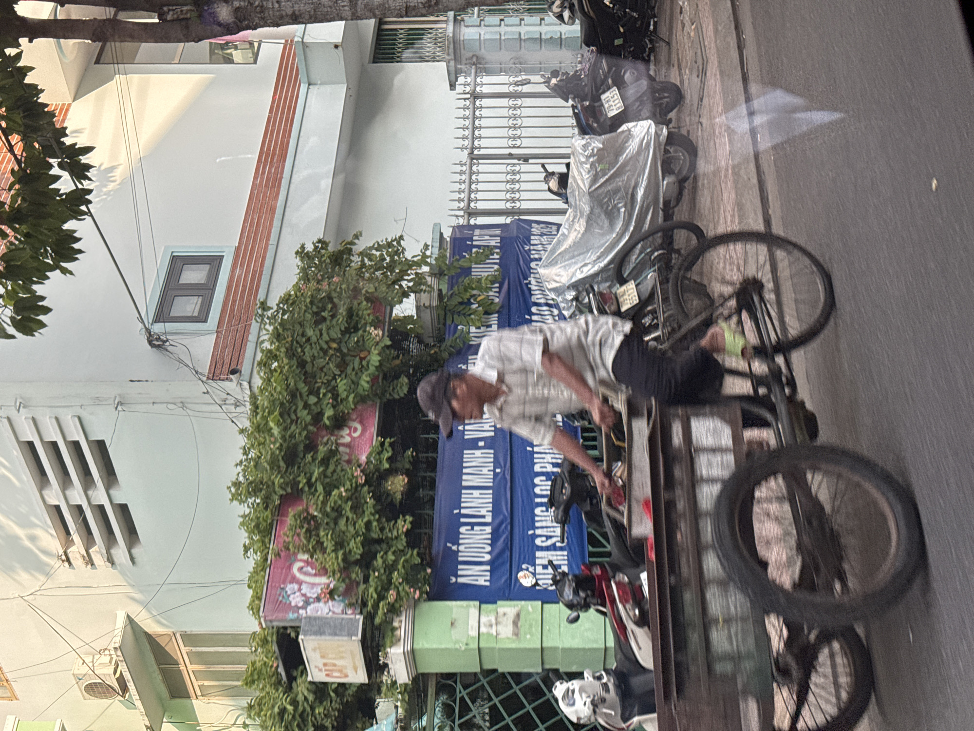 The image captures a street scene in Nha Trang, Vietnam, likely during the day, given the sunlight casting shadows. In the foreground, a person is riding a three-wheeled bicycle cart loaded with items. The rider, wearing a striped short-sleeved shirt and dark pants, is obscured from the waist up by the cart and a shadow. The cart itself is a dark-colored, open-top box with visible wheels. Behind the bicycle cart, several motorcycles are parked or are part of the traffic. One motorcycle is partially covered with a clear plastic sheet, and another has a license plate that reads "79-H1 111.53".
The background reveals a building with light-colored walls, possibly painted a pale green or blue. The building has multiple windows and what appears to be an air conditioning unit mounted on an exterior ledge. A large, dark green bush or tree obscures part of the building. A blue banner with white text in Vietnamese hangs prominently on a fence. The fence itself is decorative, made of metal bars. A sign with floral imagery and the text "CẢP" is visible above the fence. The overall impression is a typical urban street scene in Vietnam, characterized by the presence of motorcycles, street vendors, and the unique architecture.