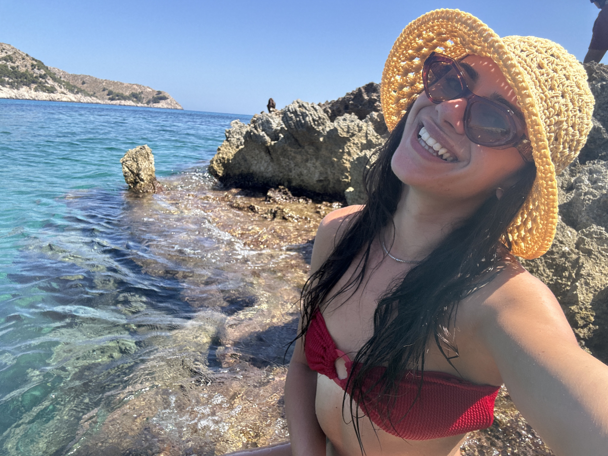A woman is captured in a close-up selfie while sitting on rocks by the sea. She is wearing a red bikini top and a yellow straw hat, with large sunglasses obscuring her eyes. Her mouth is open in a wide smile, revealing white teeth, and her long, dark hair is wet and falls around her shoulders. A delicate silver necklace adorns her neck. The background shows the clear turquoise water of the sea with rocky outcrops and a distant, hilly coastline under a bright blue sky. The sun is shining, indicating it is daytime and likely a warm day, consistent with a beach setting. The overall mood is one of joy and relaxation, suggesting the woman is enjoying her time at Cala Lliteres, Spain.