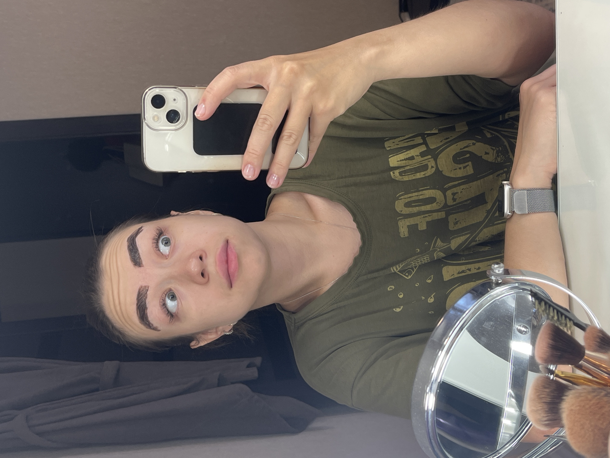 In an indoor setting, a young woman with dark hair pulled back takes a selfie in a mirror. She is wearing a green t-shirt with a graphic print. Her eyebrows appear heavily drawn, and she has a surprised expression with her eyes looking upward. To her right, she holds a white smartphone. In the foreground, a silver vanity mirror reflects a portion of the room. A collection of makeup brushes is visible in the mirror's reflection. The background shows a gray bathrobe hanging on a rack and a brown wall. The scene appears to be set in a domestic environment, likely a bathroom or bedroom. No specific landmarks or indicators place this image in Chaikovsky, Russia.