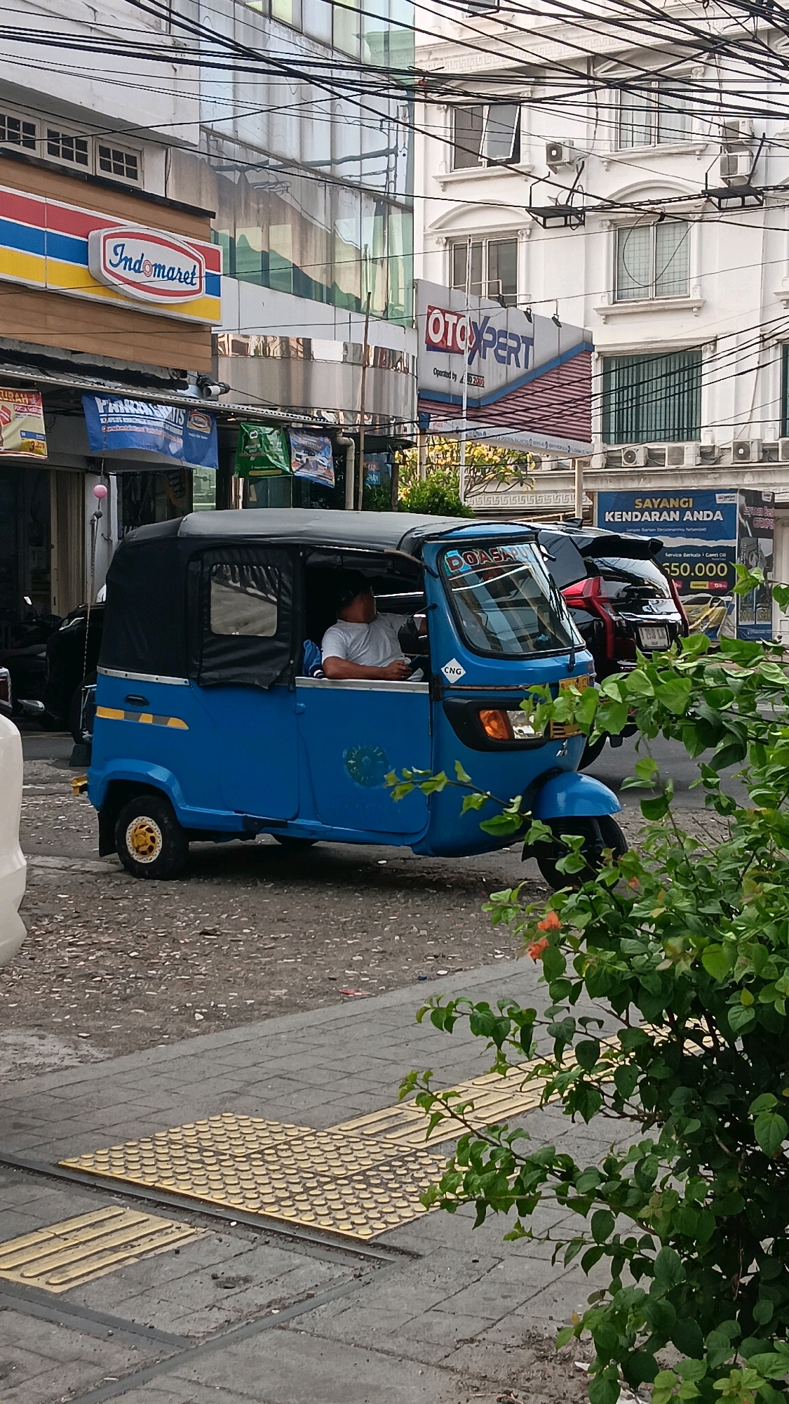 In a city street scene in South Jakarta, Indonesia, a blue auto-rickshaw is parked on the right side of the road. The auto-rickshaw has a black fabric canopy and a driver wearing a white t-shirt is seated inside. The vehicle is positioned on a gravel surface next to a sidewalk that features tactile paving. In the background, a building with a reflective glass facade is visible, displaying the "Indomaret" logo on its side. Further down the street, another building has a sign that reads "OTOXPERT". The scene is further characterized by overhead electrical wires and lush green foliage in the foreground.