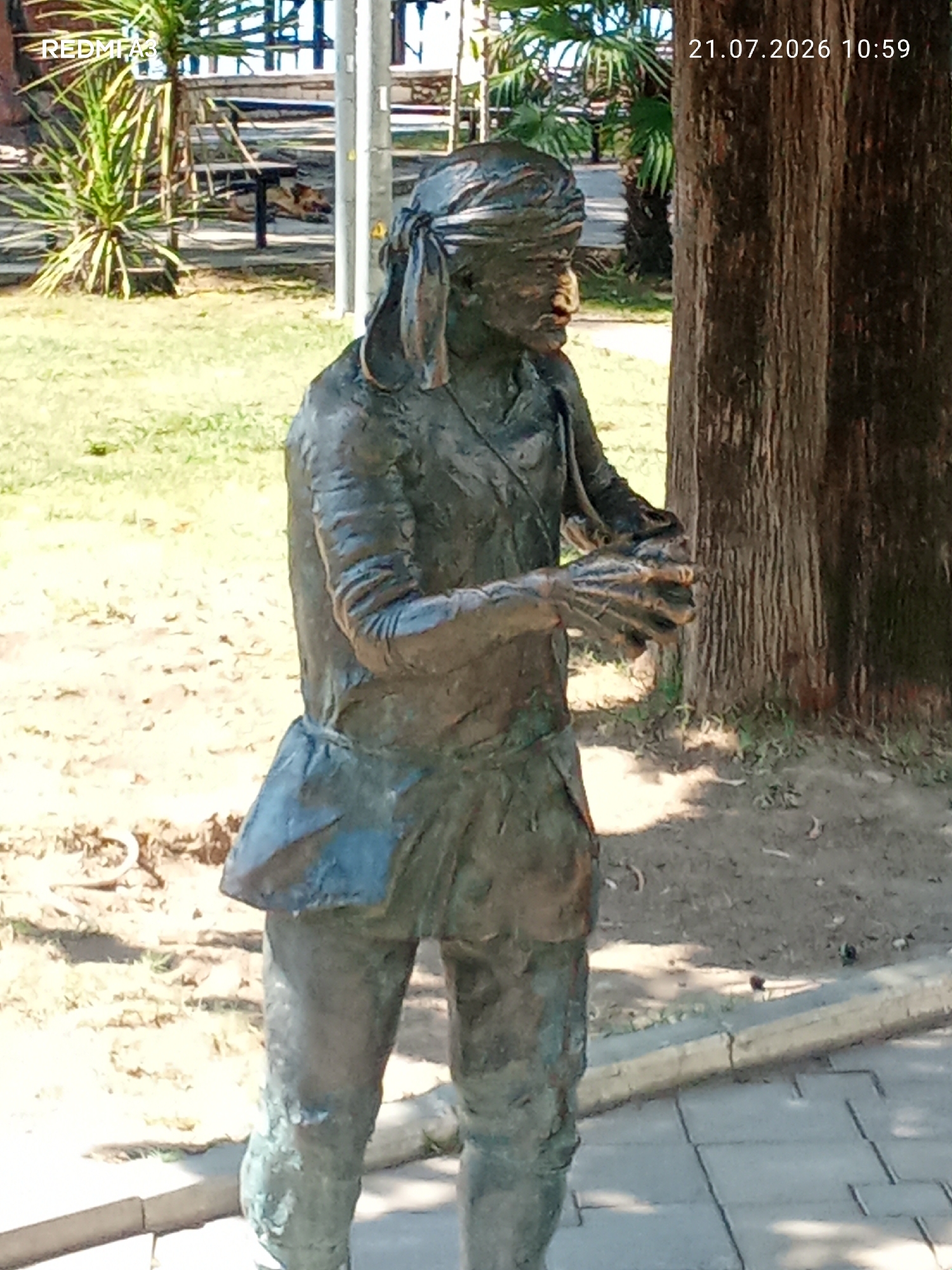 A weathered bronze statue of a man in traditional attire stands in a park-like setting in Sokhumi, Abkhazia. The statue depicts a figure with a turban-like head covering and a sash across his chest, with one hand extended forward as if holding something. Behind the statue, palm trees and other foliage are visible, along with park benches and a large tree trunk. The ground is a mix of dirt and paving stones. A date and time stamp, "21.07.2026 10:59," is present in the upper right corner of the image.