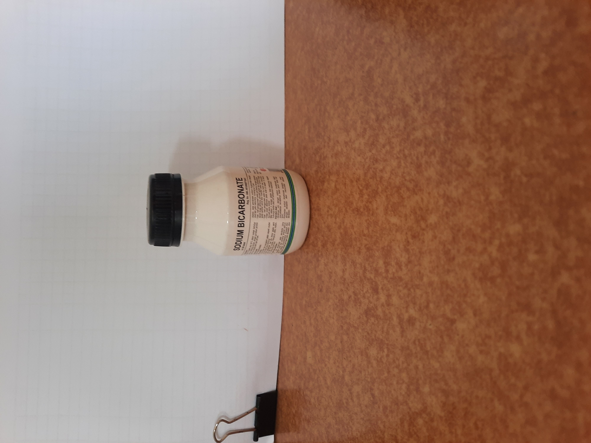 A small, clear plastic bottle of sodium bicarbonate, with a black screw-top lid, sits on a brown speckled countertop. The bottle has a white label with black text. A black binder clip is visible to the left of the bottle, attached to a white surface behind the countertop. The background is a plain white surface, possibly a wall or a piece of paper, with faint horizontal lines. The lighting is even, casting minimal shadows. The scene appears to be indoors, in a clean, uncluttered environment. There is no visible indication of the city of Lhokseumawe, Indonesia.