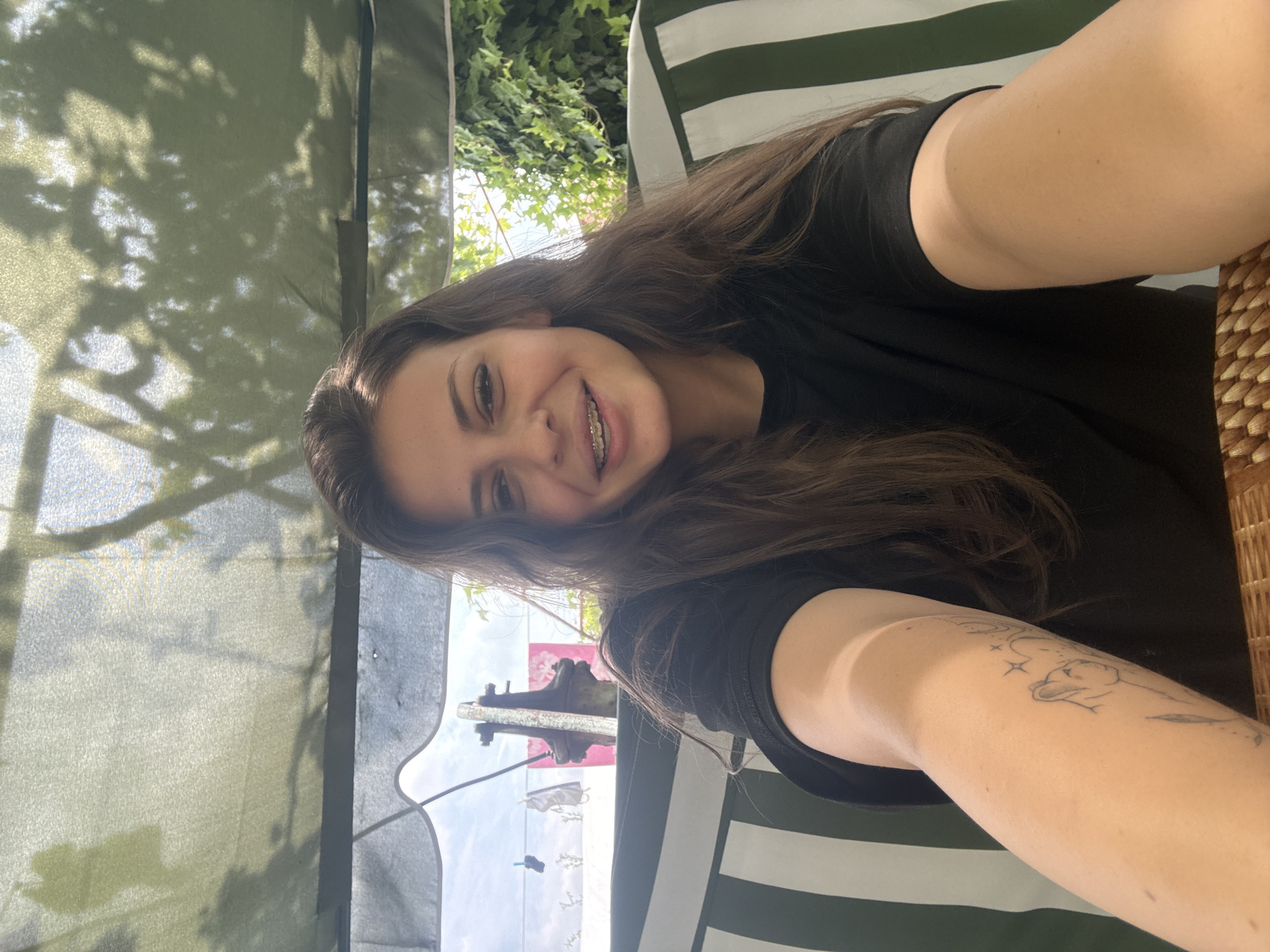 A young woman with dark brown hair and braces smiles at the camera, taking a selfie. She is wearing a black t-shirt and has a tattoo on her left arm. She is sitting on a swing with green and white striped cushions, outdoors. Behind her, there is a canopy with dappled sunlight and shadows from trees. In the background, laundry is hanging on a clothesline, and ivy covers a wall. The location is Govedare, Bulgaria.