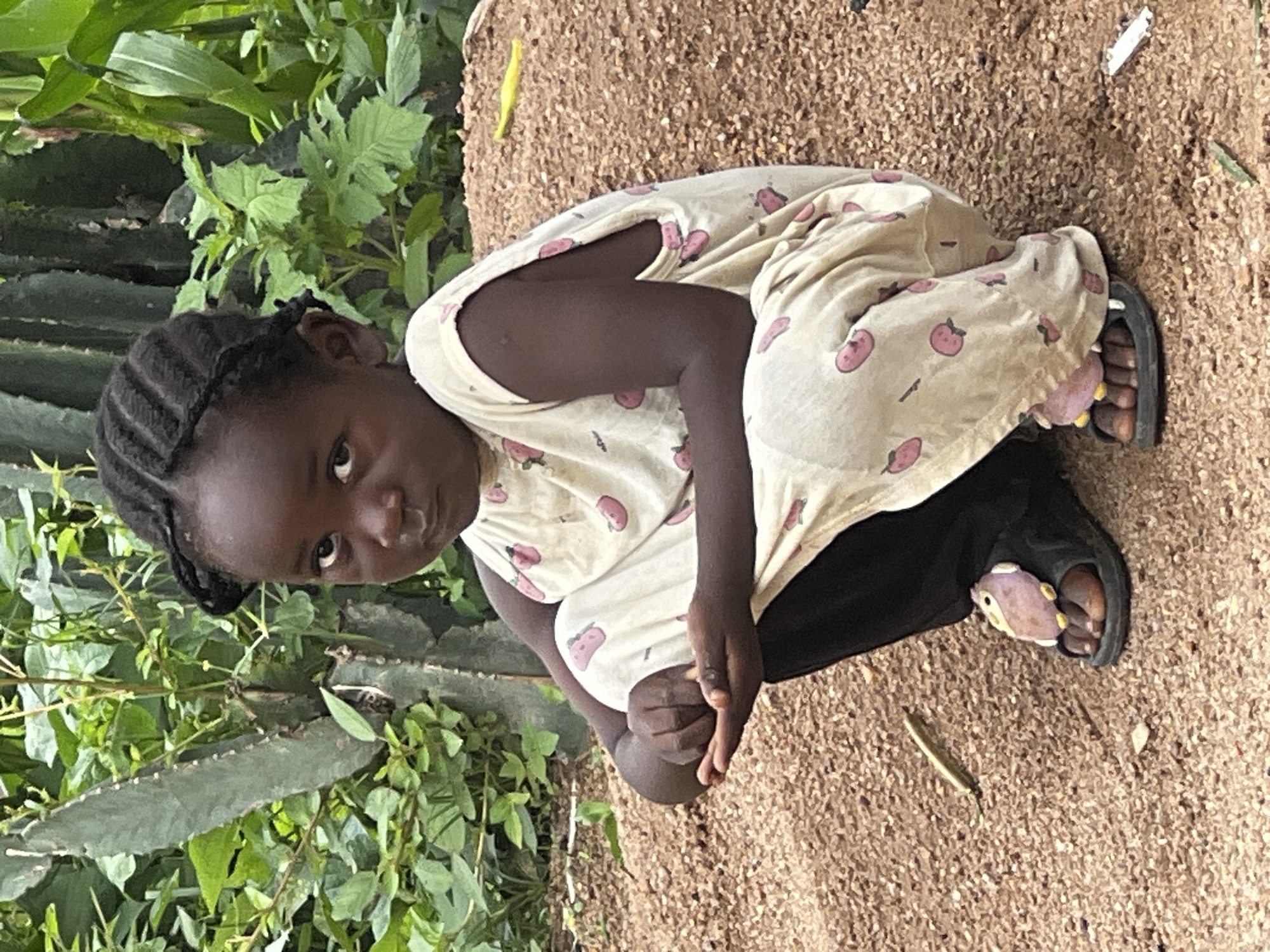 A young Black girl with braided hair and a serious expression is crouching on sandy ground. She is wearing a cream-colored pajama set with a strawberry pattern, black pants, and black sandals with owl embellishments. The background consists of green foliage, including a tall cactus, and a mound of reddish-brown dirt. The scene appears to be outdoors, possibly in a rural or semi-rural area.