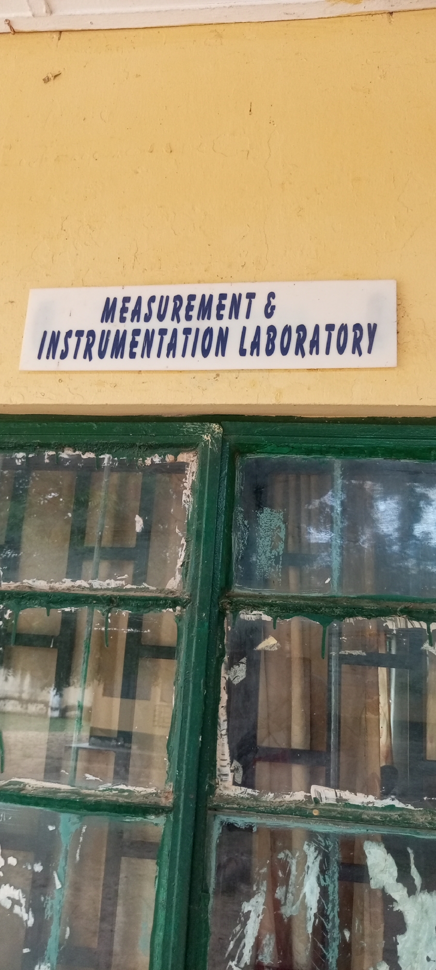 The image shows a white sign with blue text above a grimy, green-painted window frame. The sign reads "MEASUREMENT & INSTRUMENTATION LABORATORY". The window itself is divided into multiple panes by thick green mullions. The glass is dirty and smudged, with peeling white paint on the frame. Reflections in the glass obscure the view of the interior. The wall above the sign is painted a pale yellow. The image is taken from a slightly upward angle, suggesting the sign and window are on an exterior wall. The provided context places the scene in Jalingo, Nigeria.