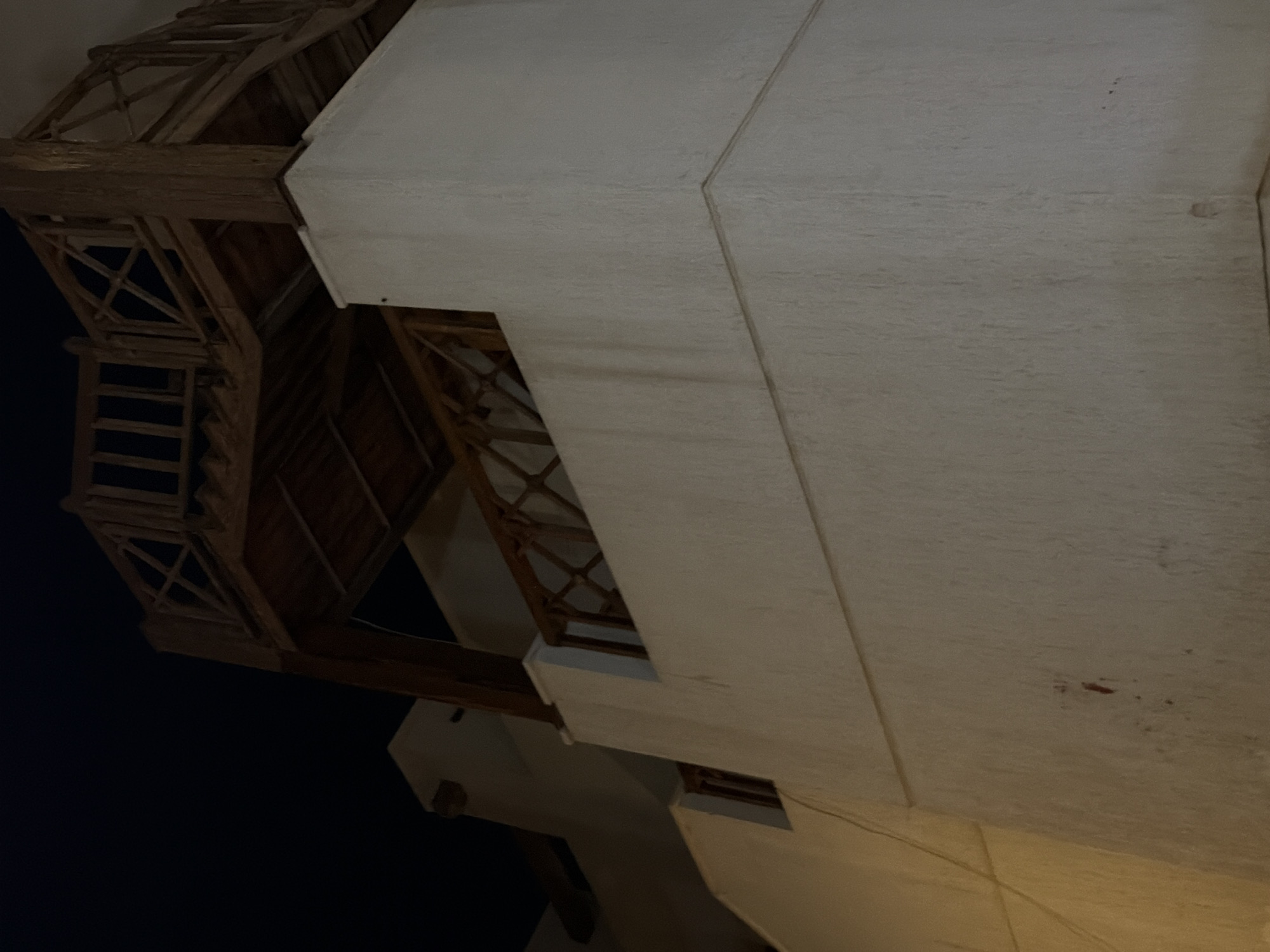 The image shows a dimly lit, external view of a white building with a wooden structure above it. The wooden structure appears to be a balcony or upper level, featuring railings with a crisscross pattern and a wooden roof. The building's wall has a textured finish, and there are some faint marks or stains on its surface. A small, dark rectangular opening is visible on the lower left portion of the building's wall. The sky in the background is dark, suggesting nighttime. The overall scene is characteristic of an outdoor architectural setting in Sharm El Sheikh, Egypt.