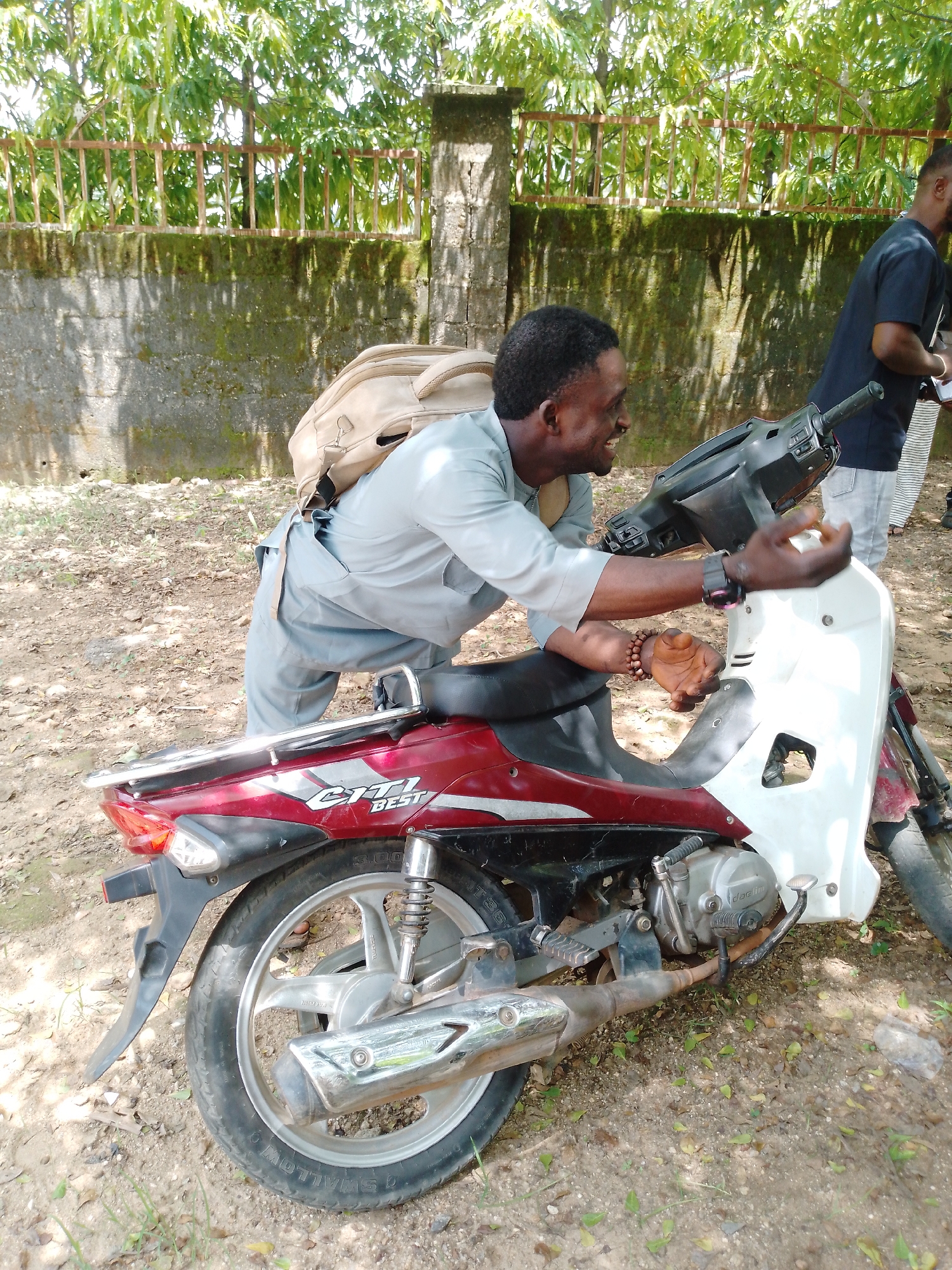 The image depicts a scene outdoors in Akwanga, Nigeria. The primary subject is a man with dark skin and short black hair, wearing a light grey top and grey trousers, with a beige backpack on his shoulders. He is leaning over a red and white motorcycle, his right arm resting on the handlebars and his left hand near the seat. He is smiling and looking to his right, appearing engaged in conversation or activity related to the motorcycle.
The motorcycle itself is a key object in the image. It is a red and white scooter-style bike with chrome accents, including a luggage rack at the back and a prominent exhaust pipe. Visible text on the motorcycle reads "CITI BEST".
Another person, a man wearing a black t-shirt and light-colored trousers, is partially visible on the right side of the frame, standing behind the motorcycle. He appears to be looking down at something in his hands, possibly a phone.
The setting is outdoors, with dry, dusty ground covered in scattered leaves and some sparse green vegetation. Behind the subjects, a moss-covered concrete wall runs horizontally, topped by a rusty metal fence. Lush green trees and foliage are visible beyond the fence, suggesting a sunny day, likely daytime given the lighting and shadows. The overall mood appears casual and perhaps social.