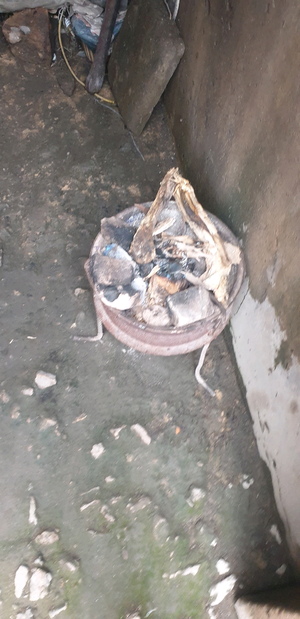 In Sabon Tasha, Nigeria, this image captures a scene within an outdoor, likely derelict, area. The ground is uneven and appears to be dirt or a similar rough surface, with scattered rocks and debris. In the foreground, a metal basin, possibly part of a makeshift stove or a discarded container, holds a collection of burnt materials. These materials include what appear to be dry vegetation, possibly wood or straw, and some darker, charred fragments. The basin itself is circular with a raised rim and shows signs of rust or wear.
To the left of the basin, a large, irregular-shaped stone rests on the ground. Behind this, a wooden pole or beam leans against other objects, including what looks like a torn piece of fabric or plastic sheeting. Yellow and white wires or cords are visible near these objects, suggesting some form of utility or discarded electrical components.
The background consists of a rough, concrete or plastered wall, which appears weathered and possibly damp in places. The overall impression is one of disuse and neglect.