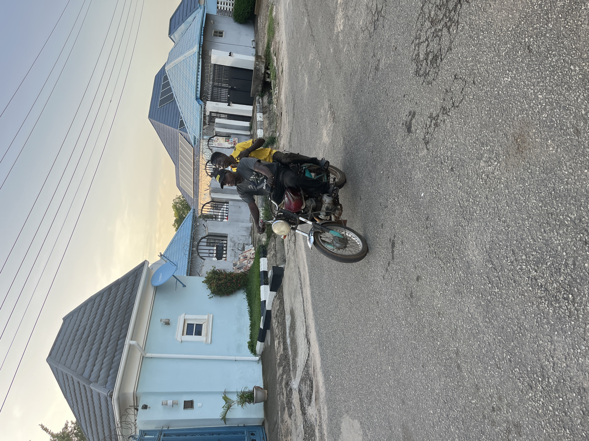 Two young men ride a motorcycle on an asphalt road lined with pastel blue houses in what appears to be a residential area in Abuja, Nigeria. The houses have varying roof designs, some with solar panels and satellite dishes. The sky is a pale gradient of blue and yellow, suggesting it may be early morning or late afternoon. Power lines are visible overhead. The road surface is worn with visible repairs and patches. The motorcycle is red and black. The rider wears a gray t-shirt and a black cap. The passenger wears a yellow shirt.