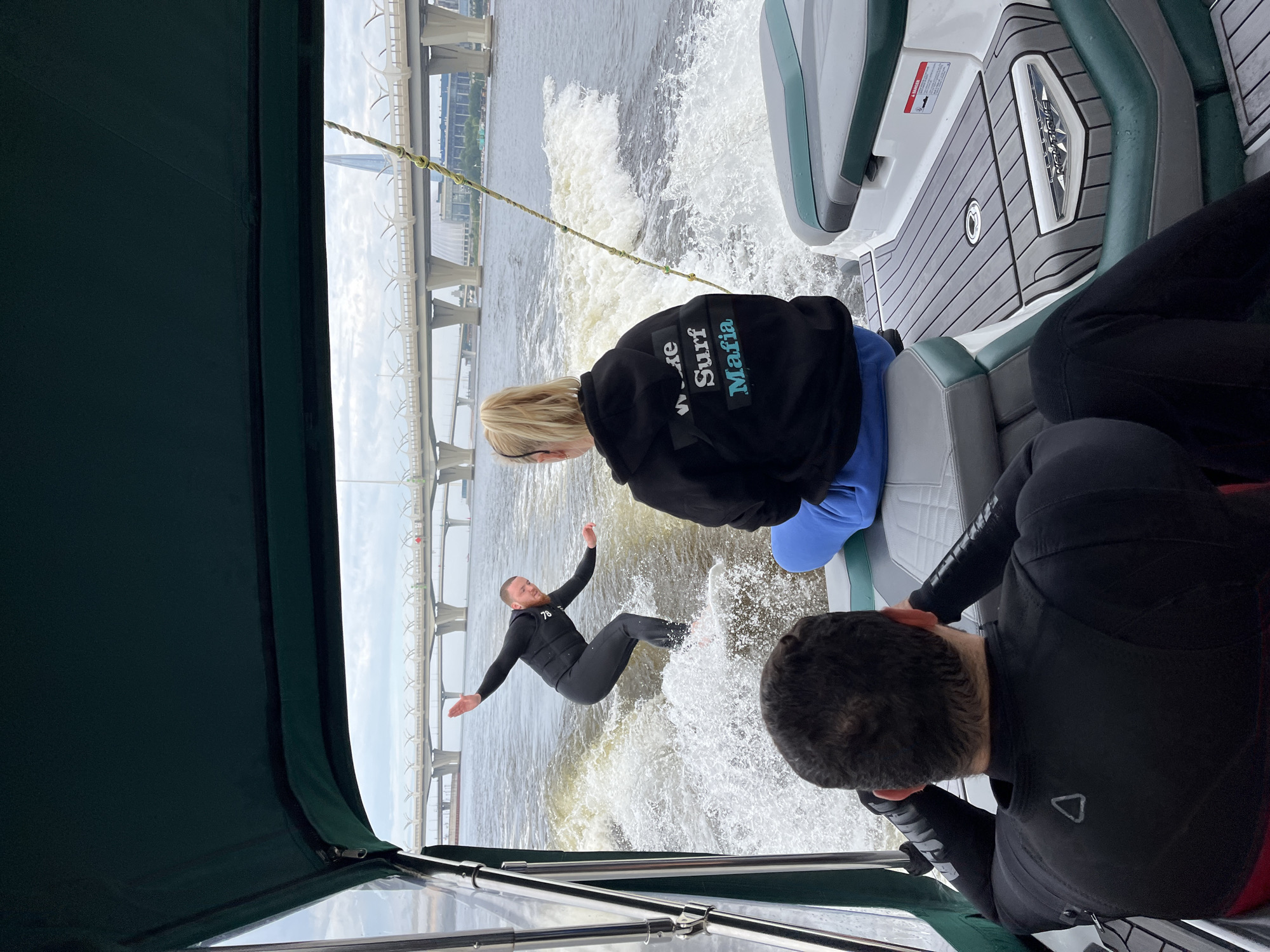 The image depicts a wake surfing activity on the Neva River in Saint Petersburg, Russia, with a distinctive bridge structure in the background. In the foreground, two individuals are seated on a boat. The person on the left, facing away from the camera, is wearing a black wetsuit. The person on the right, also facing away, is wearing a black hoodie with "Wake Surf Mafia" printed on the back in white and blue lettering, and blue sweatpants. In the mid-ground, a man in a black wetsuit is actively wake surfing on a white board, carving through the water and creating a significant wake. The Neva River is visible, with choppy water and spray from the surfing activity. In the background, a large, modern bridge with white arches spans the river. The sky is overcast, suggesting a cloudy day. The distinctive architecture of the bridge, particularly its multiple levels and the way it curves, is a recognizable landmark in Saint Petersburg, likely the Western High-Speed Diameter (WHSD) bridge.