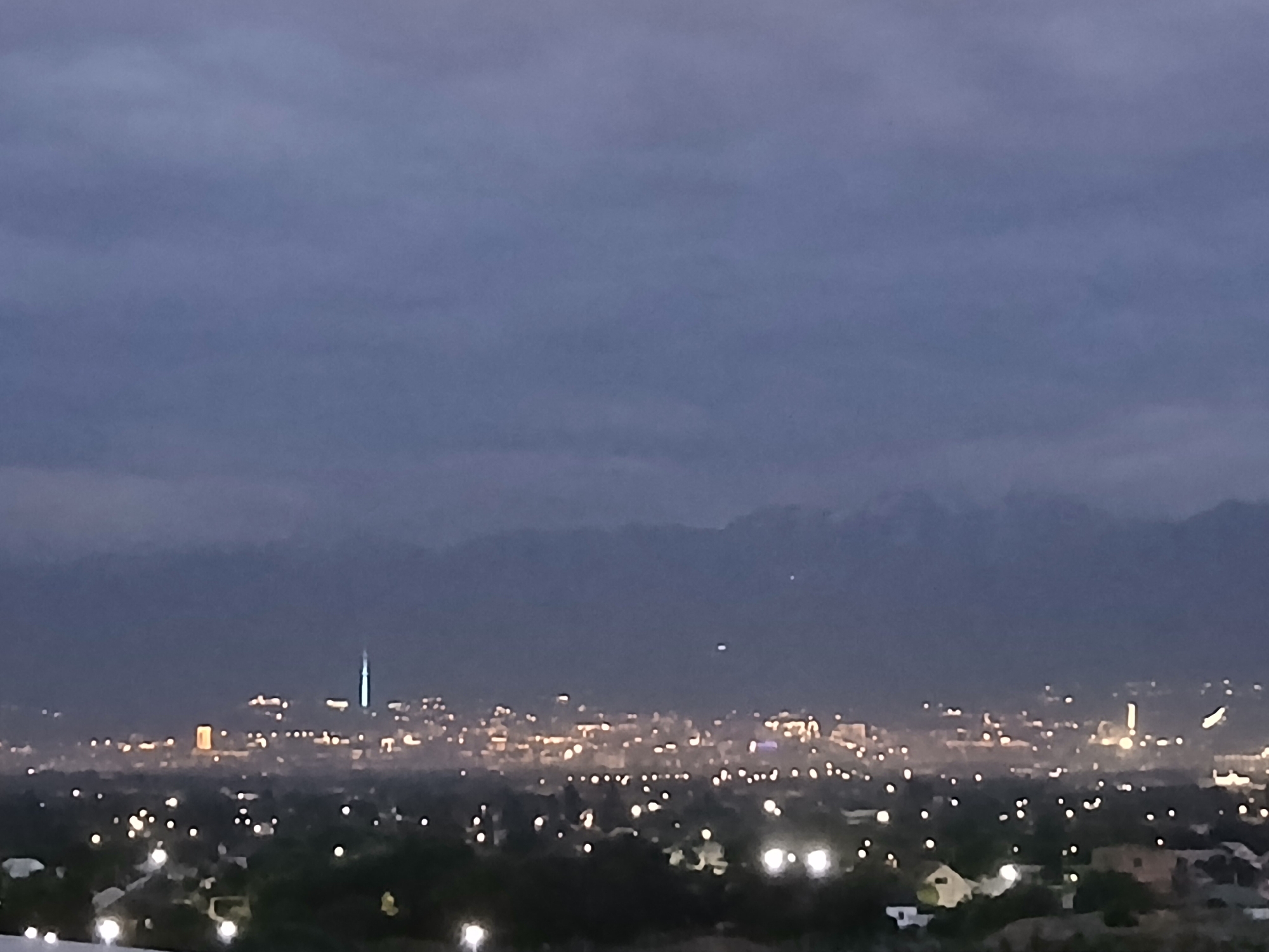The image depicts a blurred, twilight panorama of a city with scattered lights. In the foreground, dark silhouettes of trees and a few illuminated structures are visible. The middle ground shows a denser concentration of city lights, suggesting a developed urban area. A prominent, tall, illuminated structure with a pointed top, possibly a tower or spire, stands out in the middle-left of the cityscape. In the background, a range of dark mountains is silhouetted against a dark, cloudy sky. The overall atmosphere is dim and hazy, indicative of either early morning or late evening conditions.